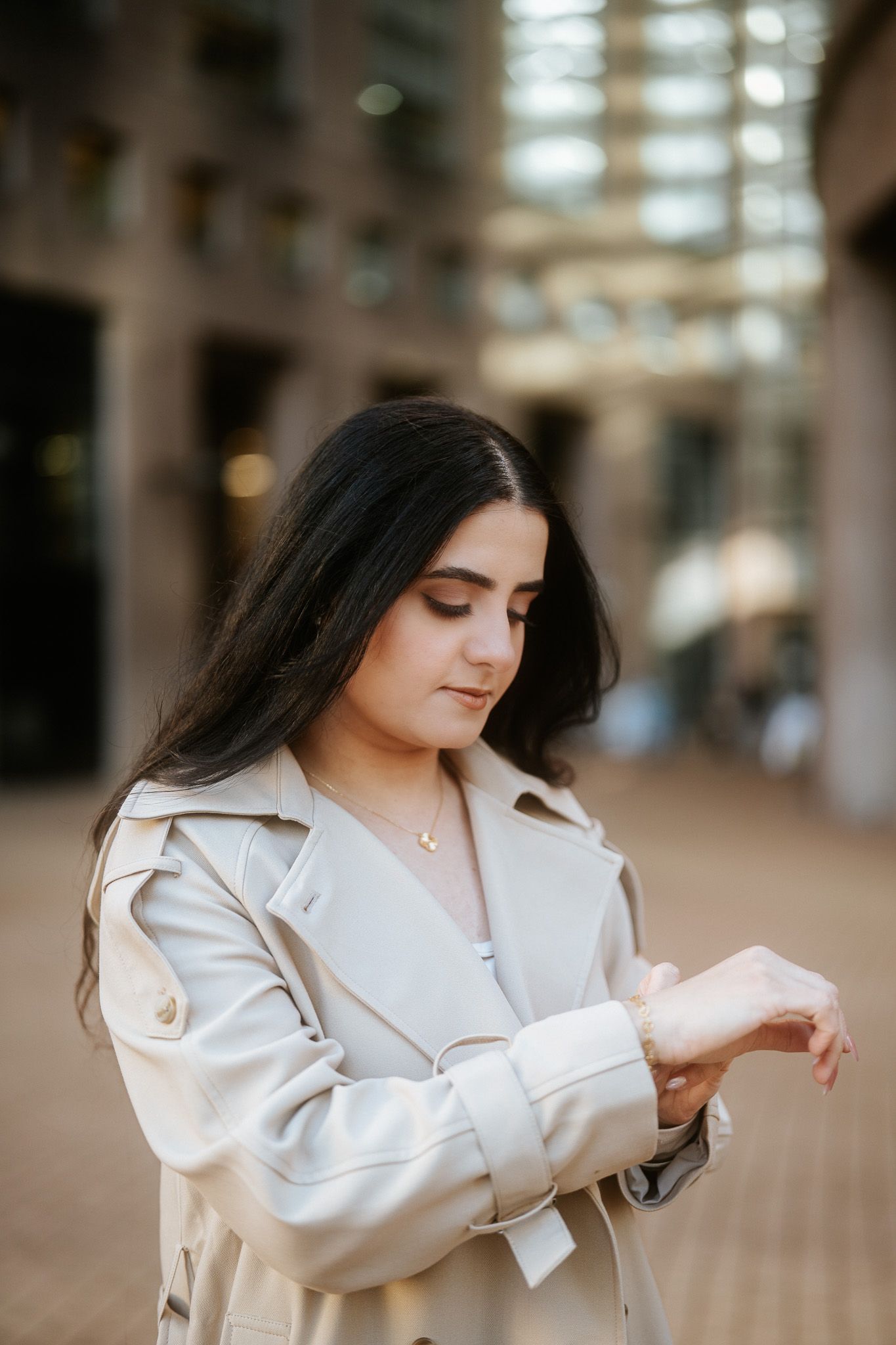 Person in white blazer stands in soft lighting while looking down at their sleeve in a contemplative pose.
