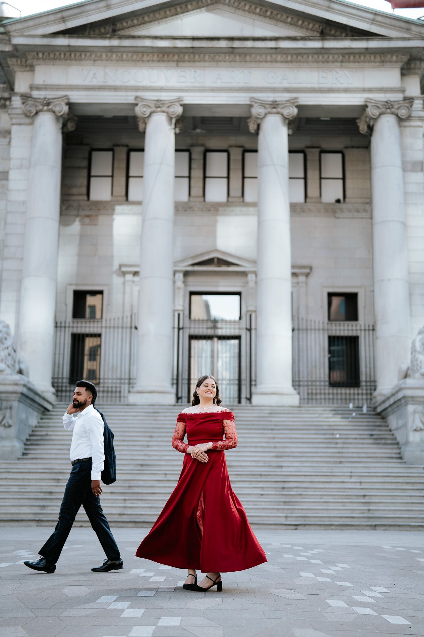Woman in elegant red off-shoulder gown twirling before a neoclassical building with tall white columns.