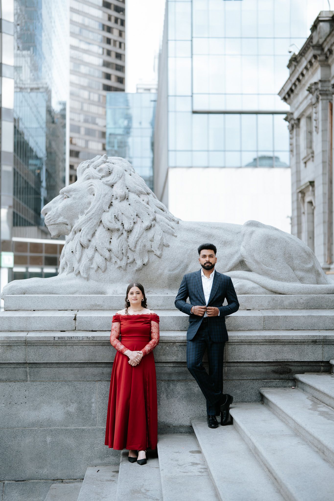 Couple posing by a stone lion sculpture, woman in red gown and man in dark suit, urban architecture backdrop.