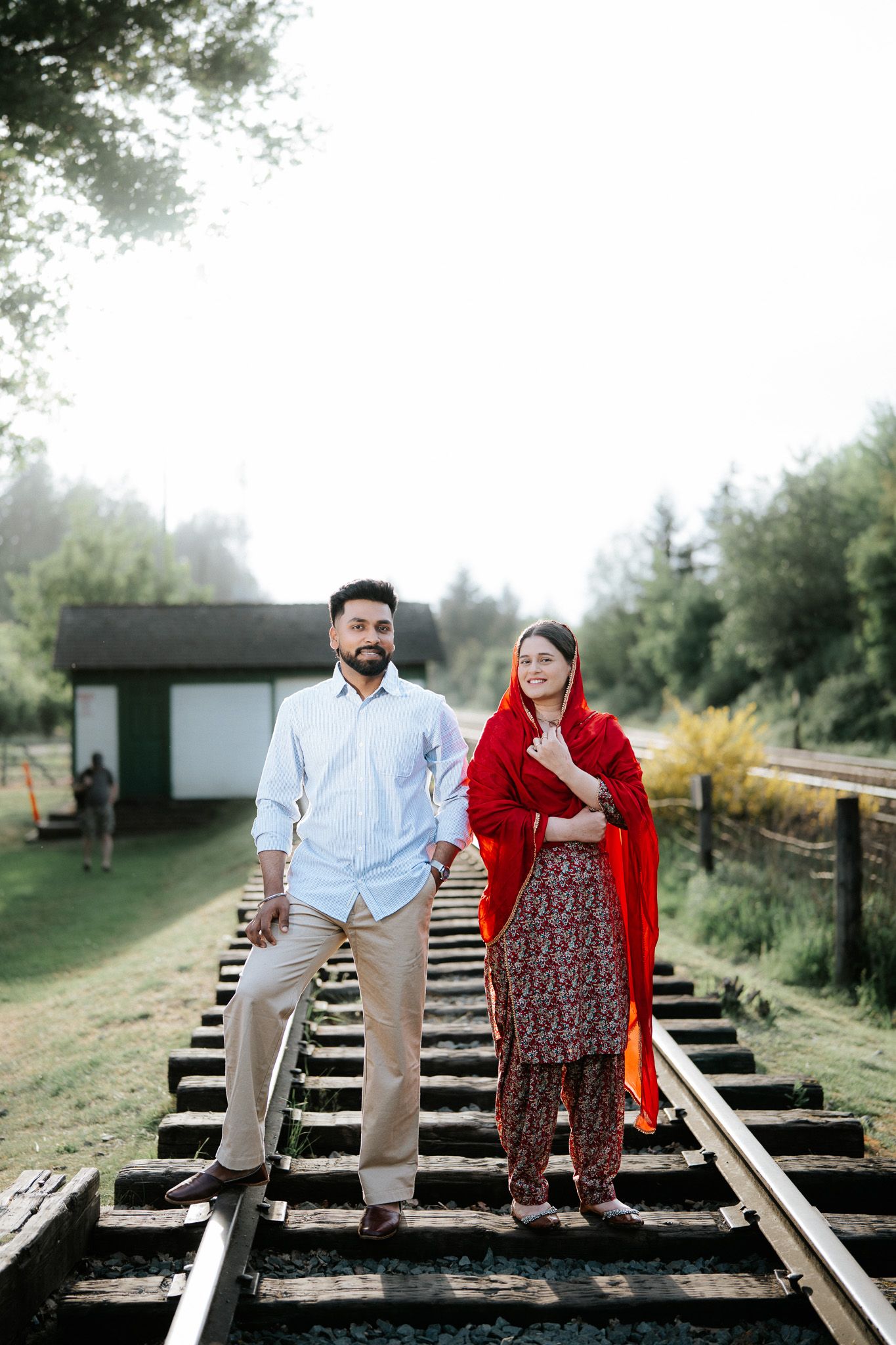 Couple standing on railway tracks, woman in red salwar kameez and man in light blue shirt, surrounded by lush greenery.