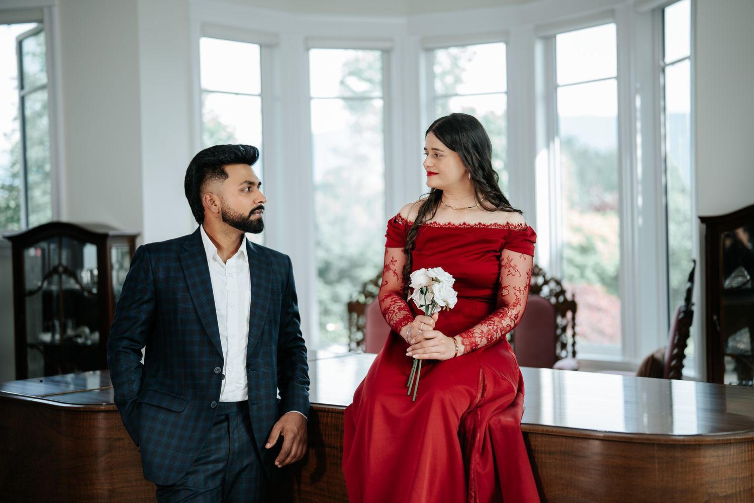 Couple standing by grand piano near bay windows, woman in red off-shoulder lace gown holding white rose bouquet.
