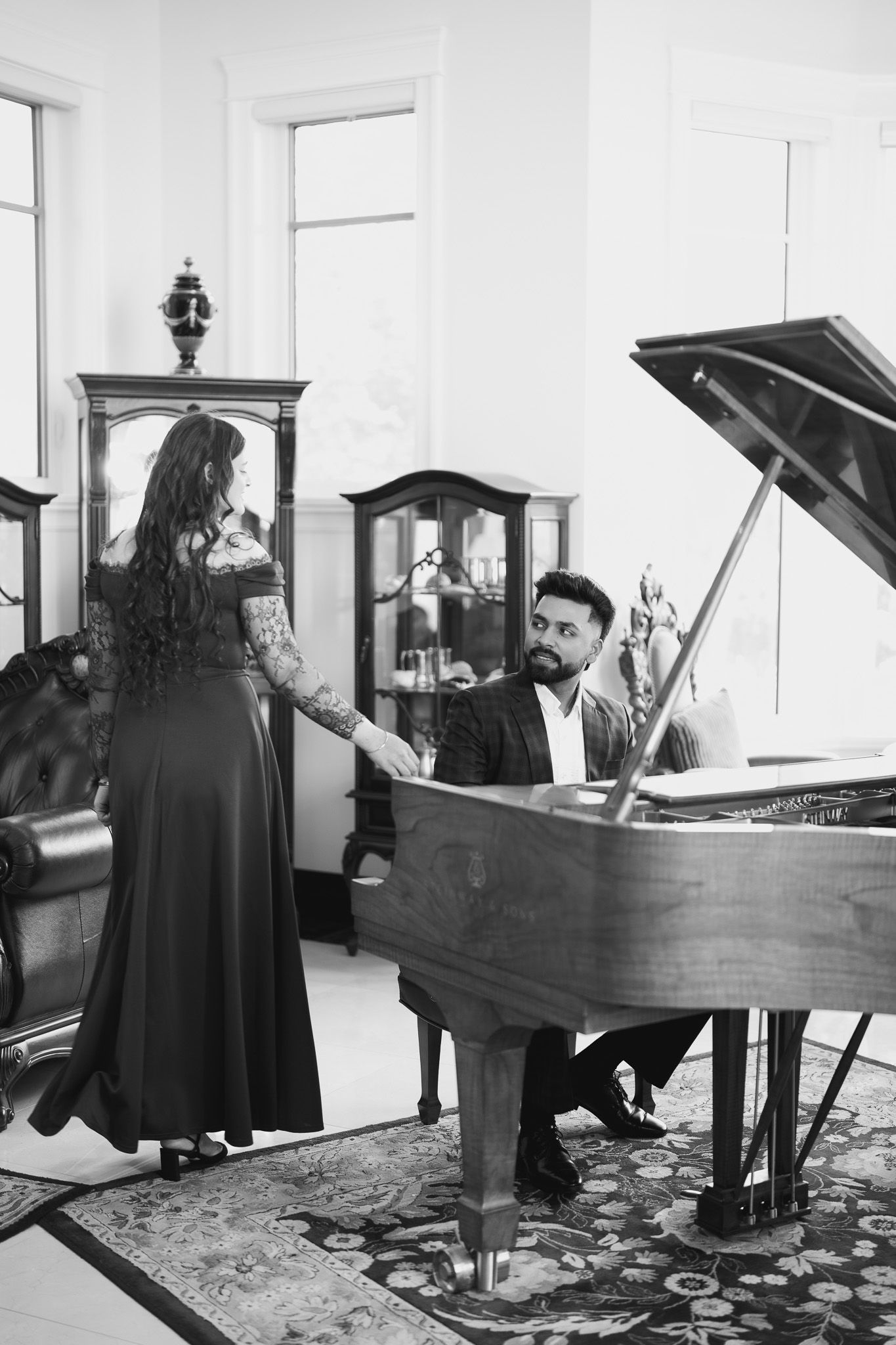 Elegant black and white photo of a bearded man playing grand piano while a tattooed woman in evening gown stands beside him.