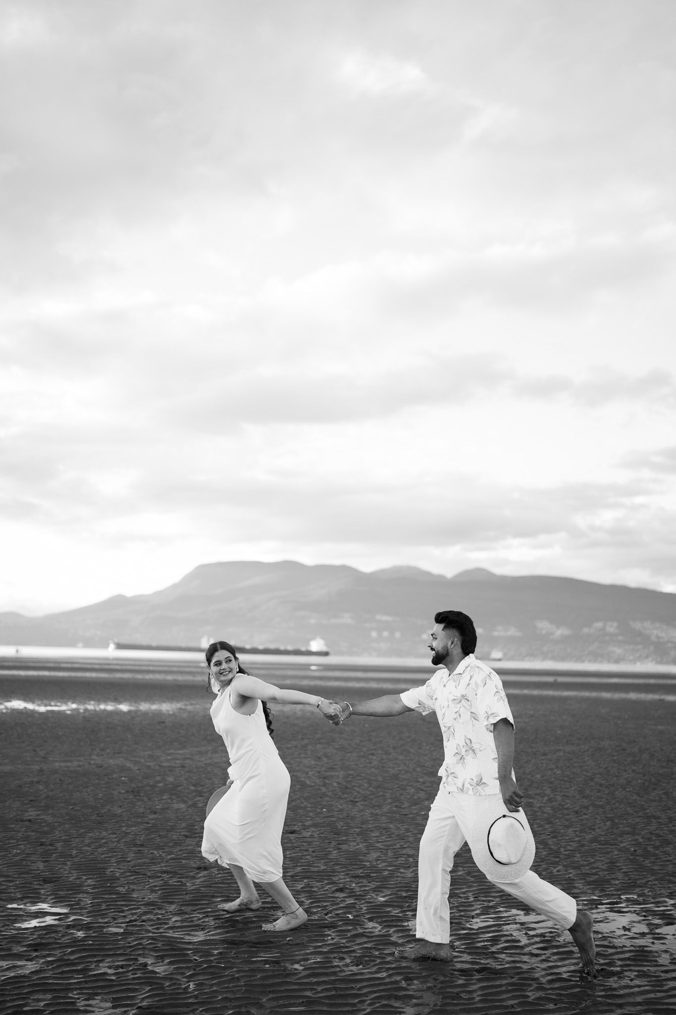 A joyful couple runs along a beach in black and white, holding hands with mountains in the background.
