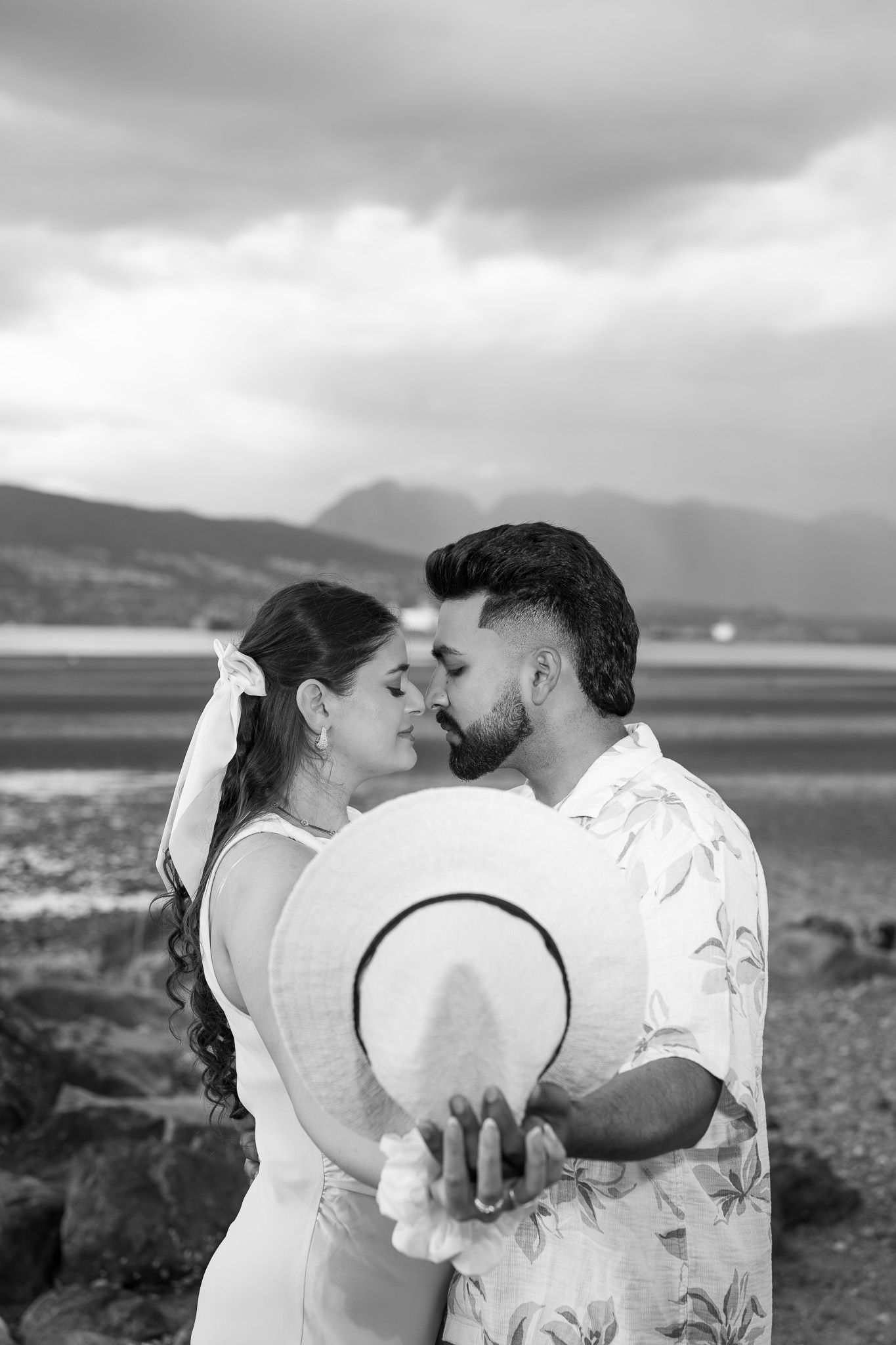 Black and white photo of couple almost kissing on beach, holding hat between them, mountains in background.