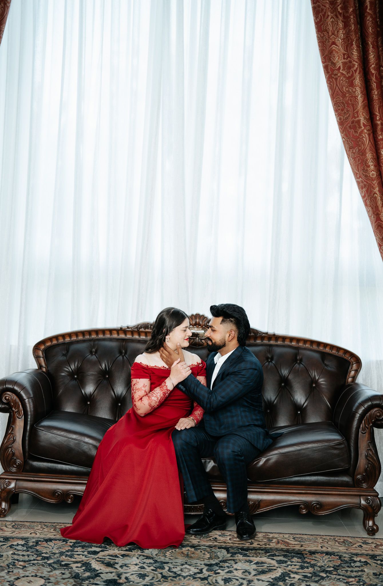 Romantic couple in formal attire sitting close on a leather sofa, woman in red lace dress, man in dark suit.