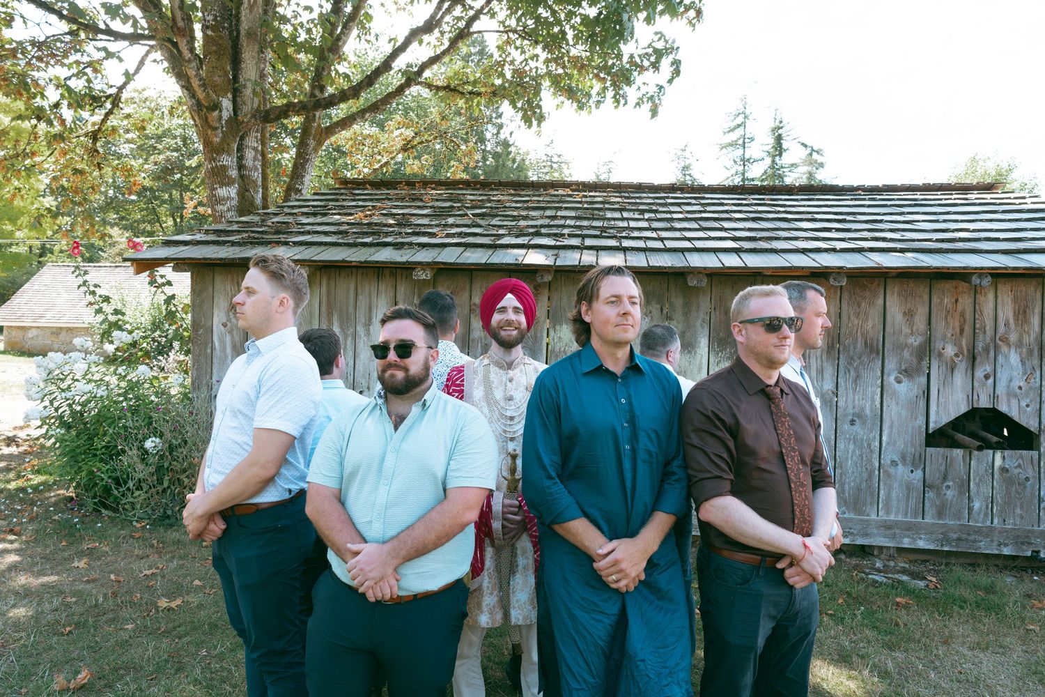 Five men in formal attire stand outdoors near a wooden shed surrounded by trees.