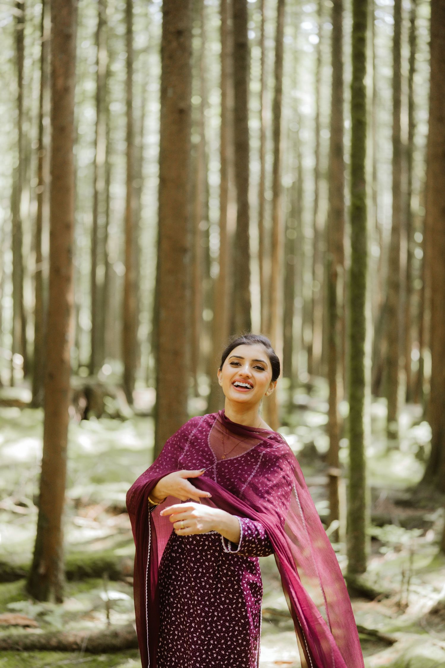 Smiling South Asian woman in burgundy salwar kameez twirling her dupatta in a tall pine forest.