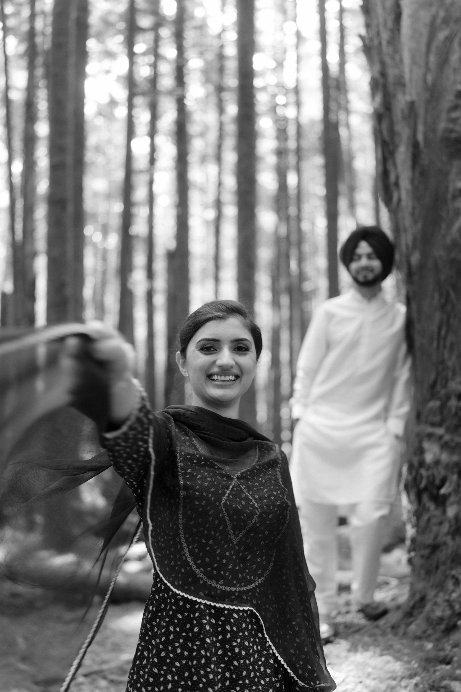 Black and white photo of a smiling South Asian woman in salwar suit twirling her dupatta in a forest.