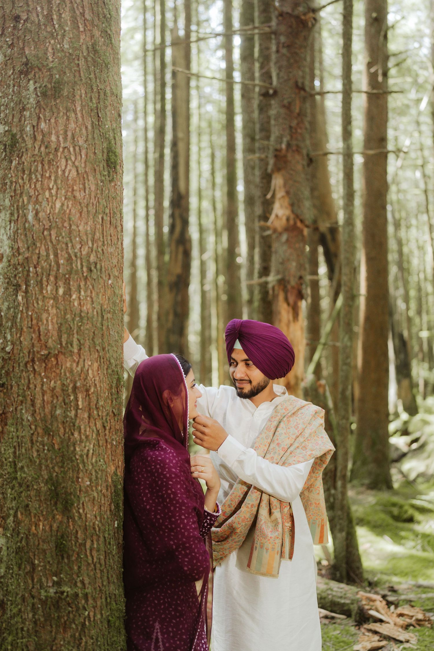 Sikh couple sharing tender moment by a tree in forest, man in purple turban smiling at woman in matching purple traditional dress.