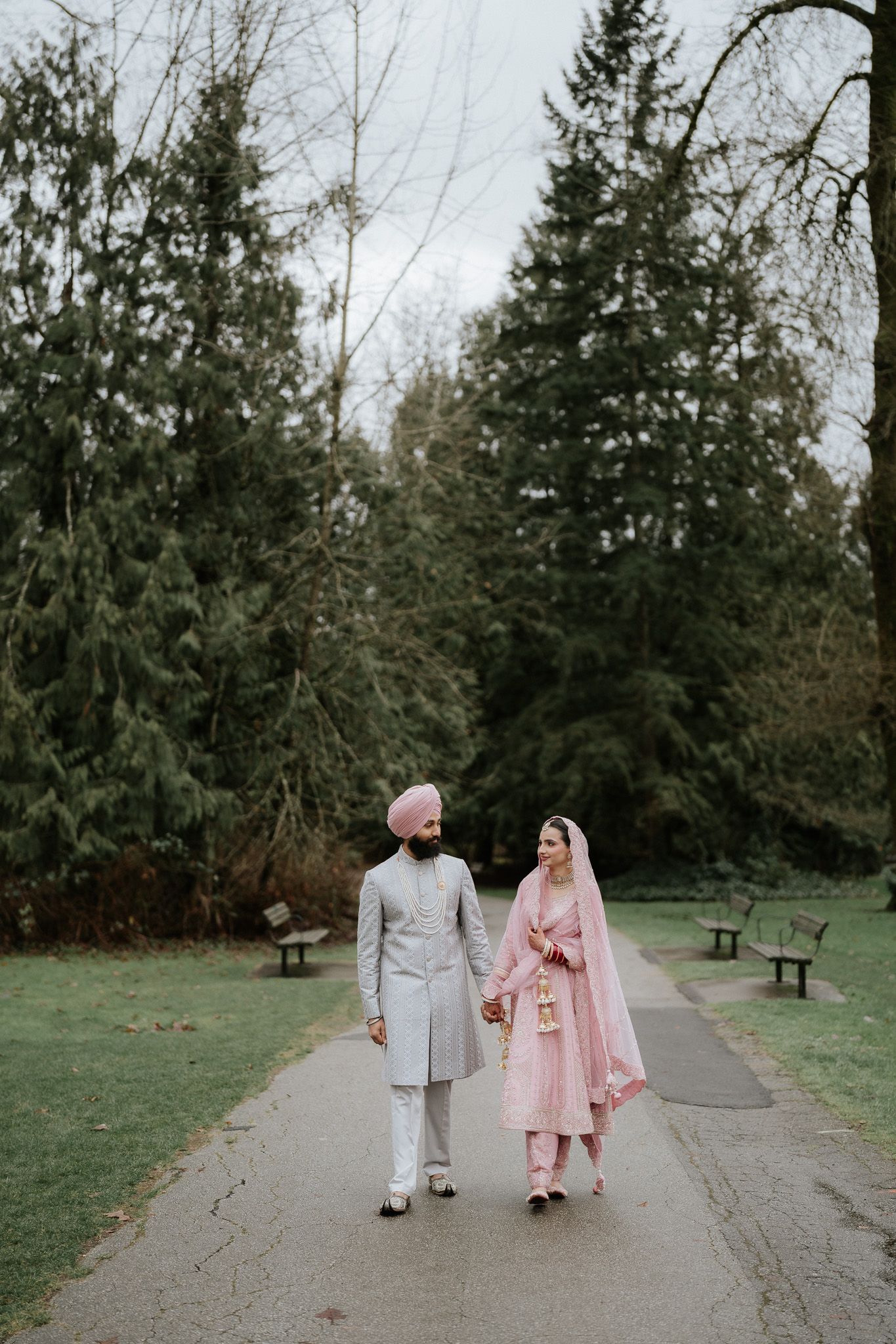 A couple in traditional wedding attire walks along a tree-lined path in a park, wearing pink and gray formal clothing.