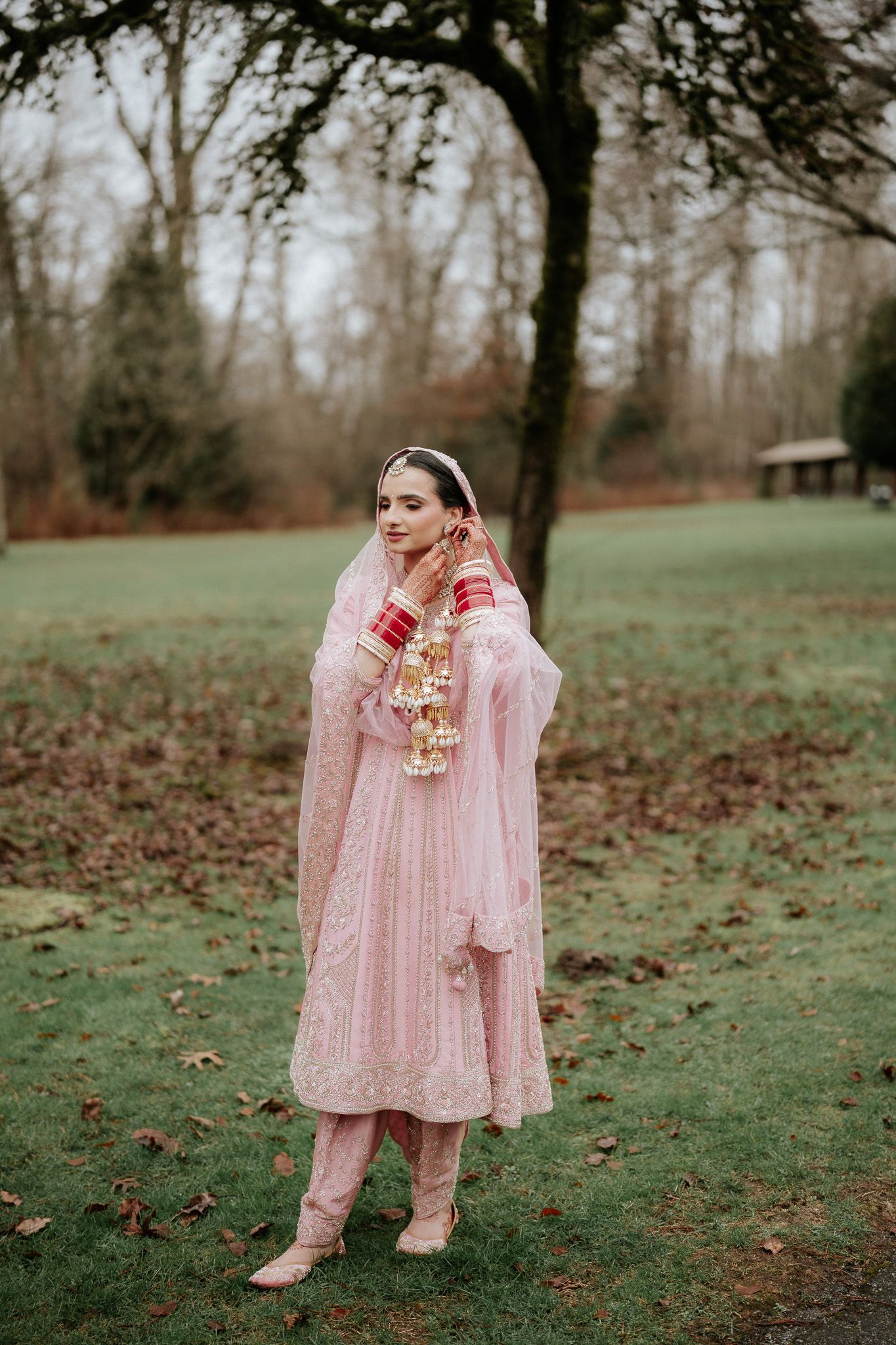 A person in a pink traditional South Asian wedding outfit stands in a misty garden setting with fallen autumn leaves.
