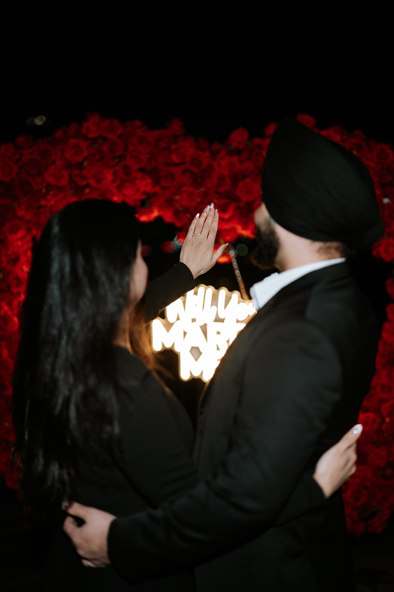 A couple in dark formal attire share a romantic moment in front of a glowing light against a deep red background.