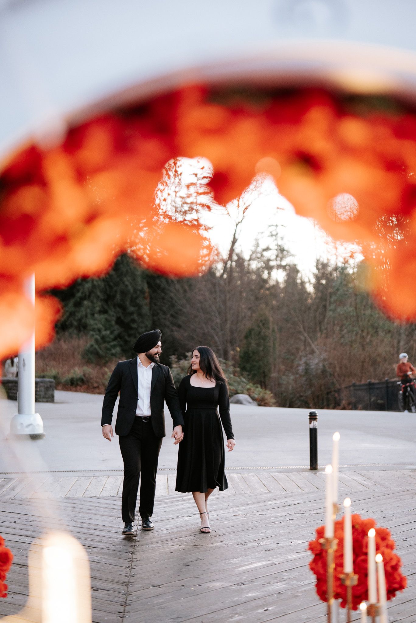 A couple walks together on a wooden deck surrounded by red floral decorations at sunset.