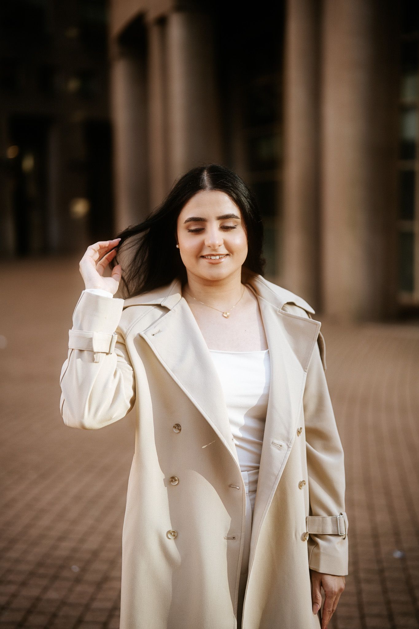 A person wearing a stylish cream-colored trench coat and white top stands outdoors with a joyful expression.
