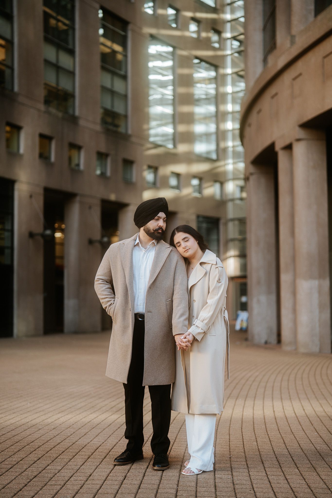 A couple in formal attire shares a romantic moment in an urban architectural setting with curved walls and columns.