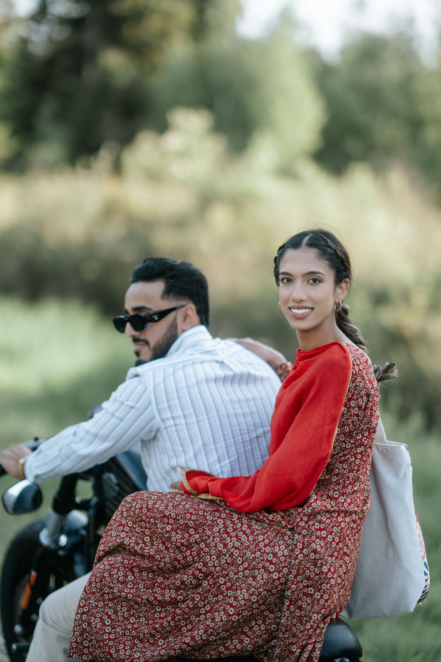 A couple riding together on a motorcycle, with the passenger wearing a red sweater and floral dress in a scenic outdoor setting.