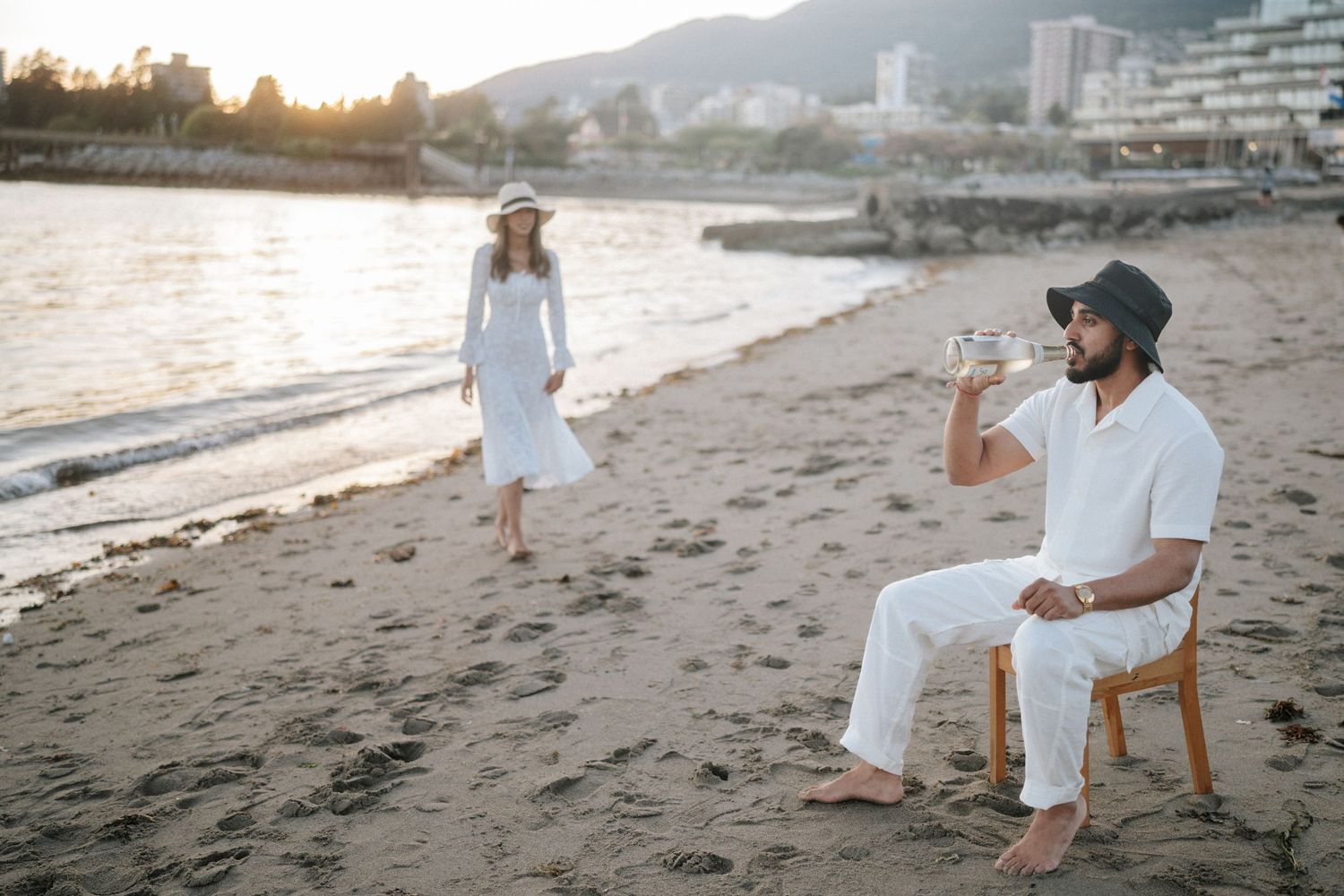 A person in white clothing sits on a beach chair at sunset with mountains and buildings in the background.