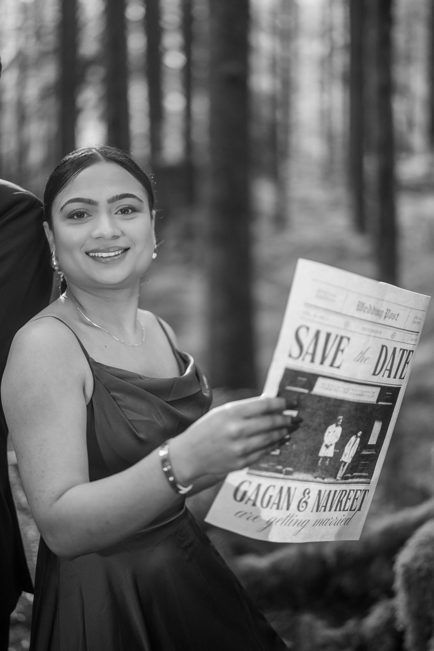 A woman in a red dress holds up a save-the-date newspaper announcement in a forest.