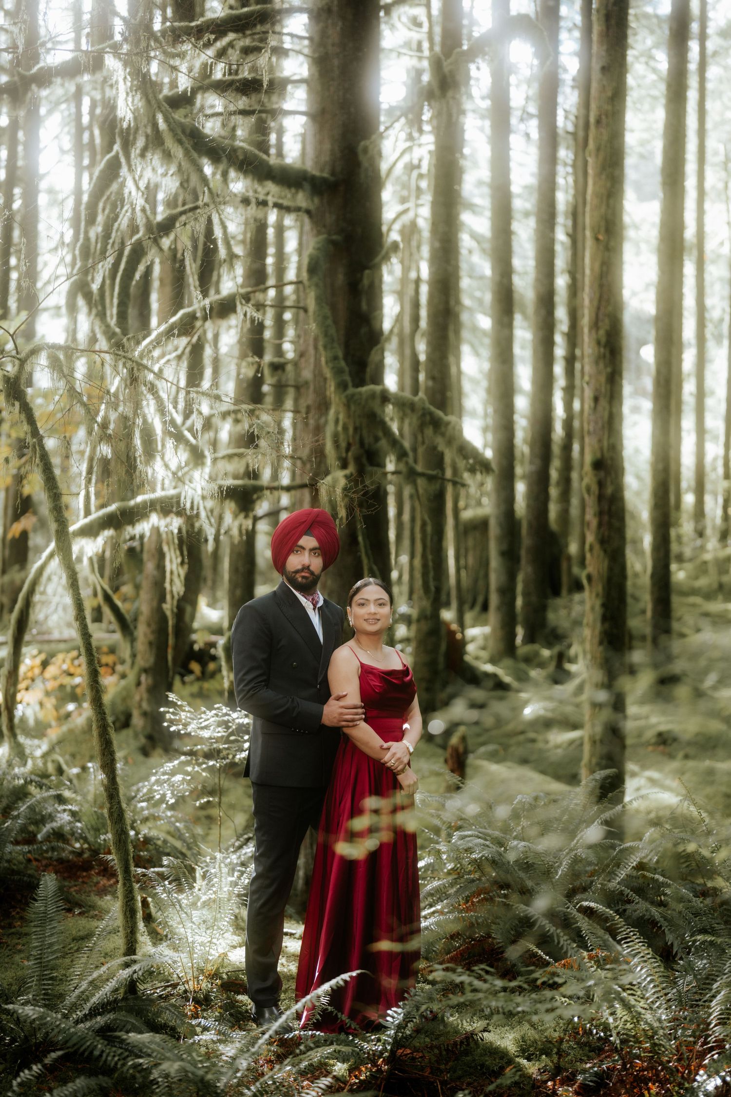 A couple in formal attire embraces among ferns in a misty forest setting.