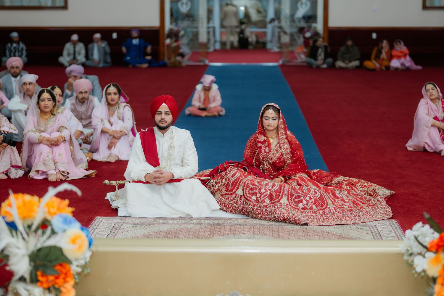 Traditional Sikh wedding ceremony with guests seated on floor in a Gurdwara with red and white decor.