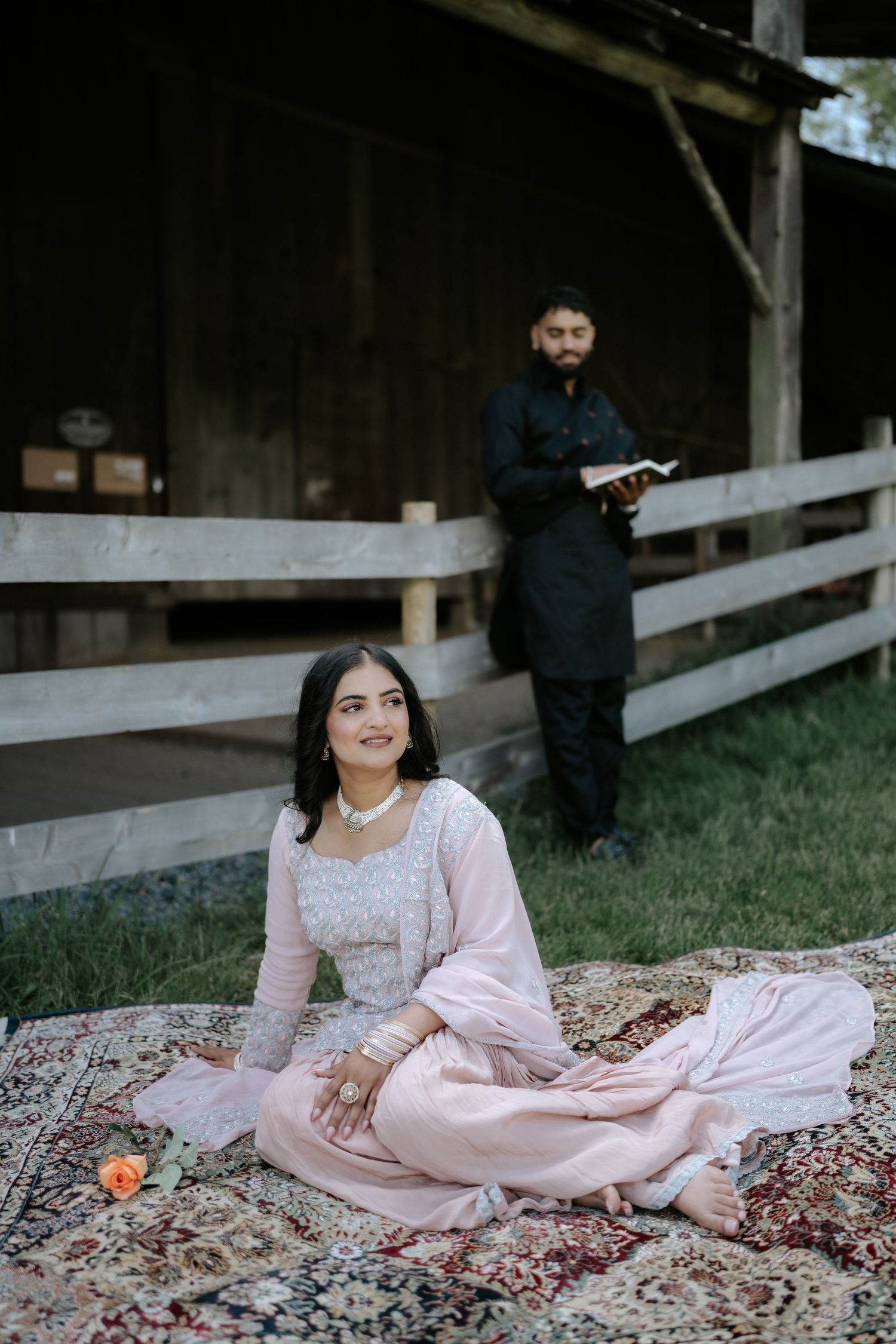 A person in pink traditional attire sits on an ornate rug in front of a wooden structure.