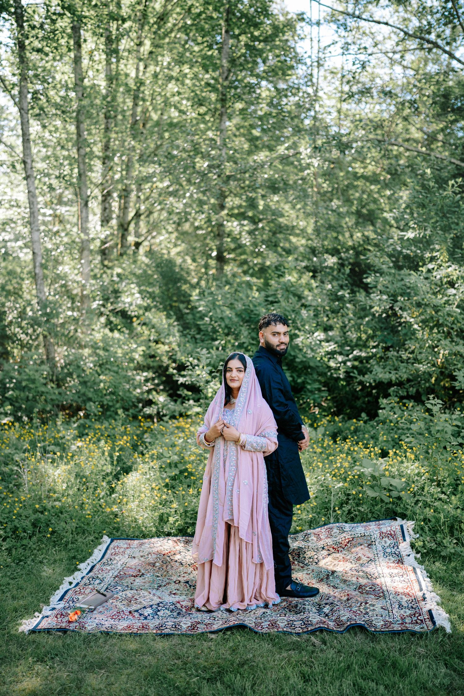 A couple sits on a patterned blanket in a lush forest setting during a romantic outdoor photoshoot.