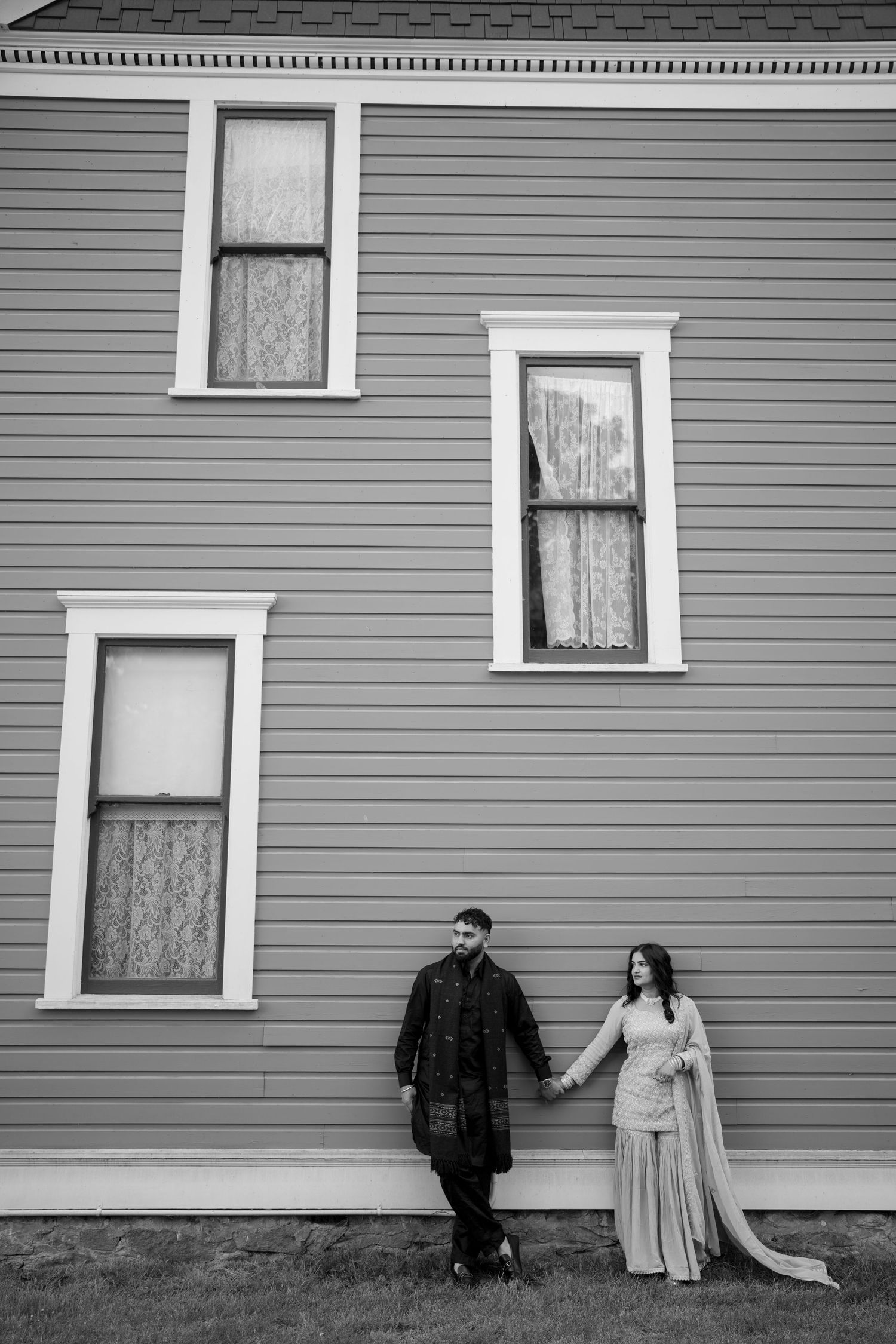 A couple poses artistically against a gray clapboard house with multiple windows.