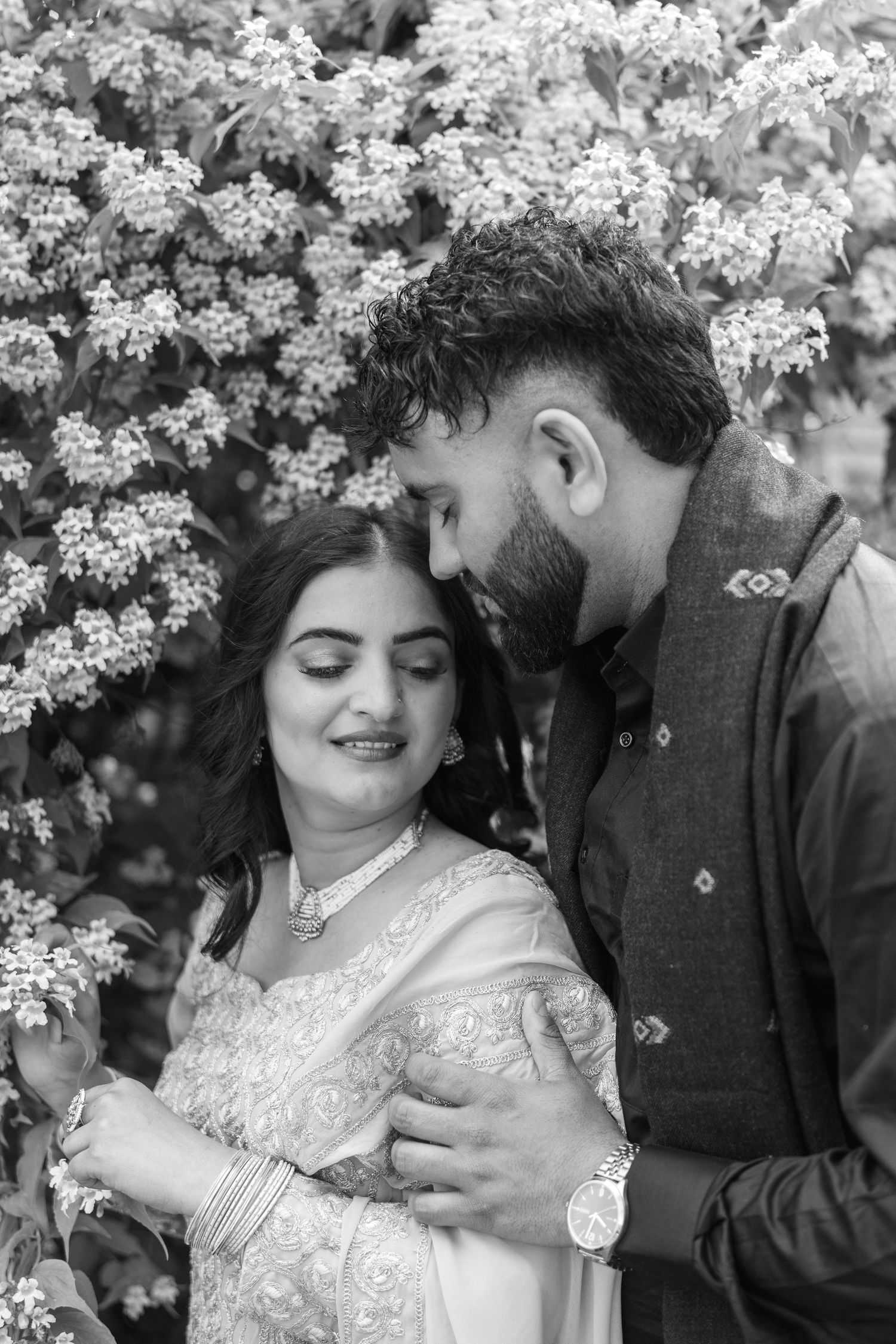 A couple shares intimate moments under blooming white flowers in black and white photography.