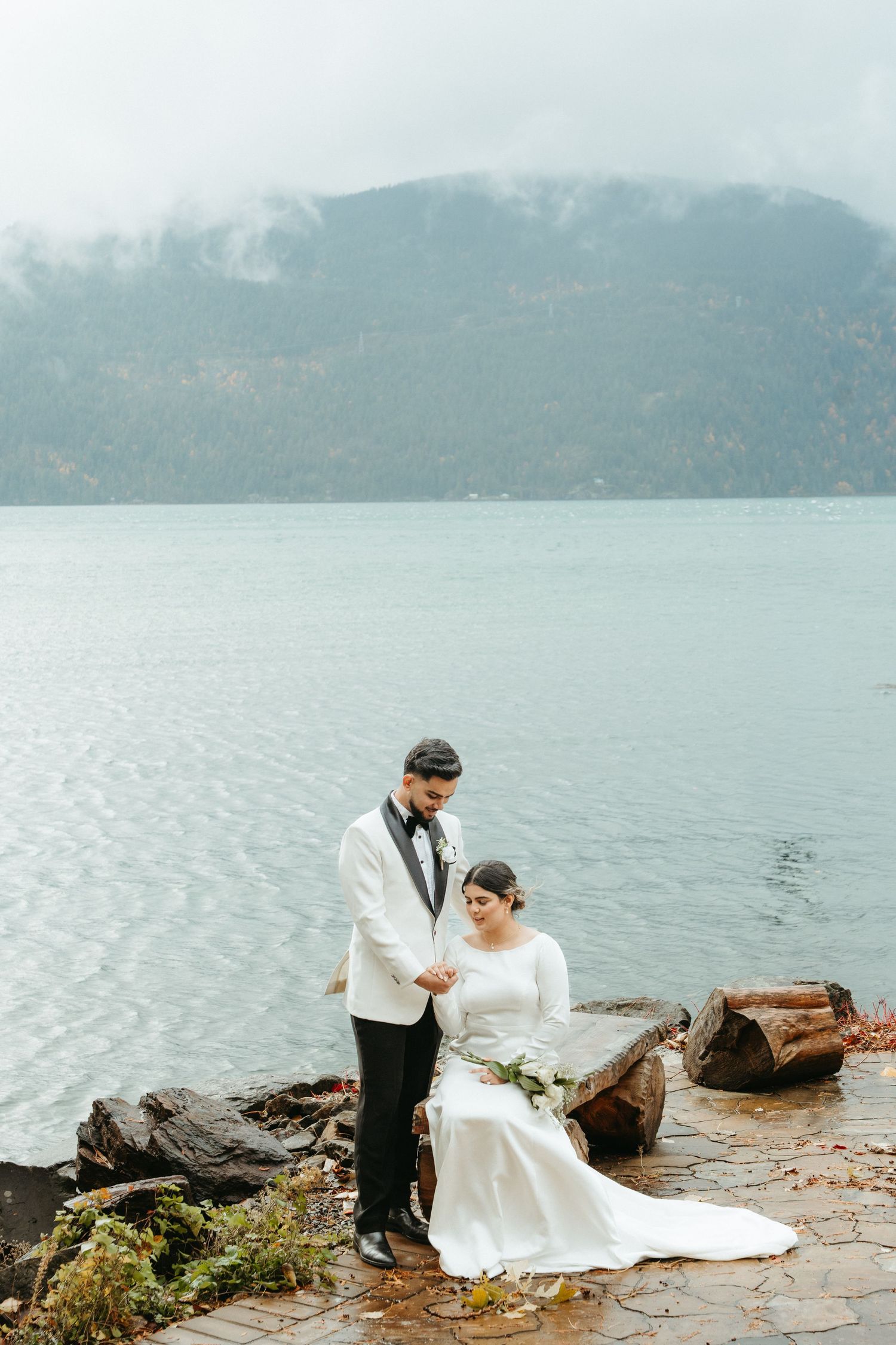 A couple in wedding attire poses by a misty lake with mountains in the background, creating a romantic atmosphere.