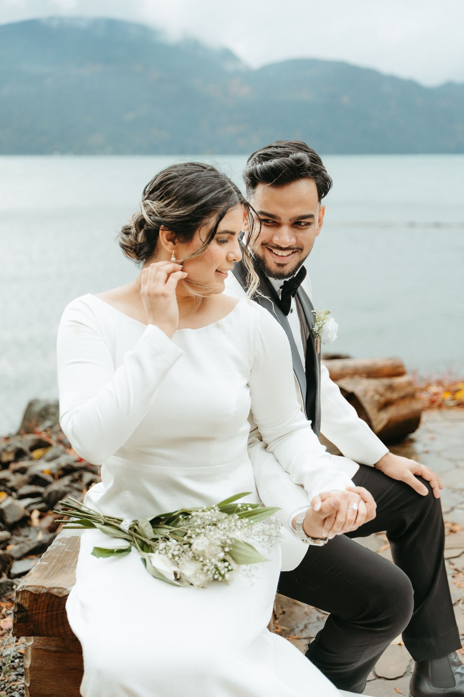 A couple shares intimate moments on a rocky beach with mountains and water in the background.