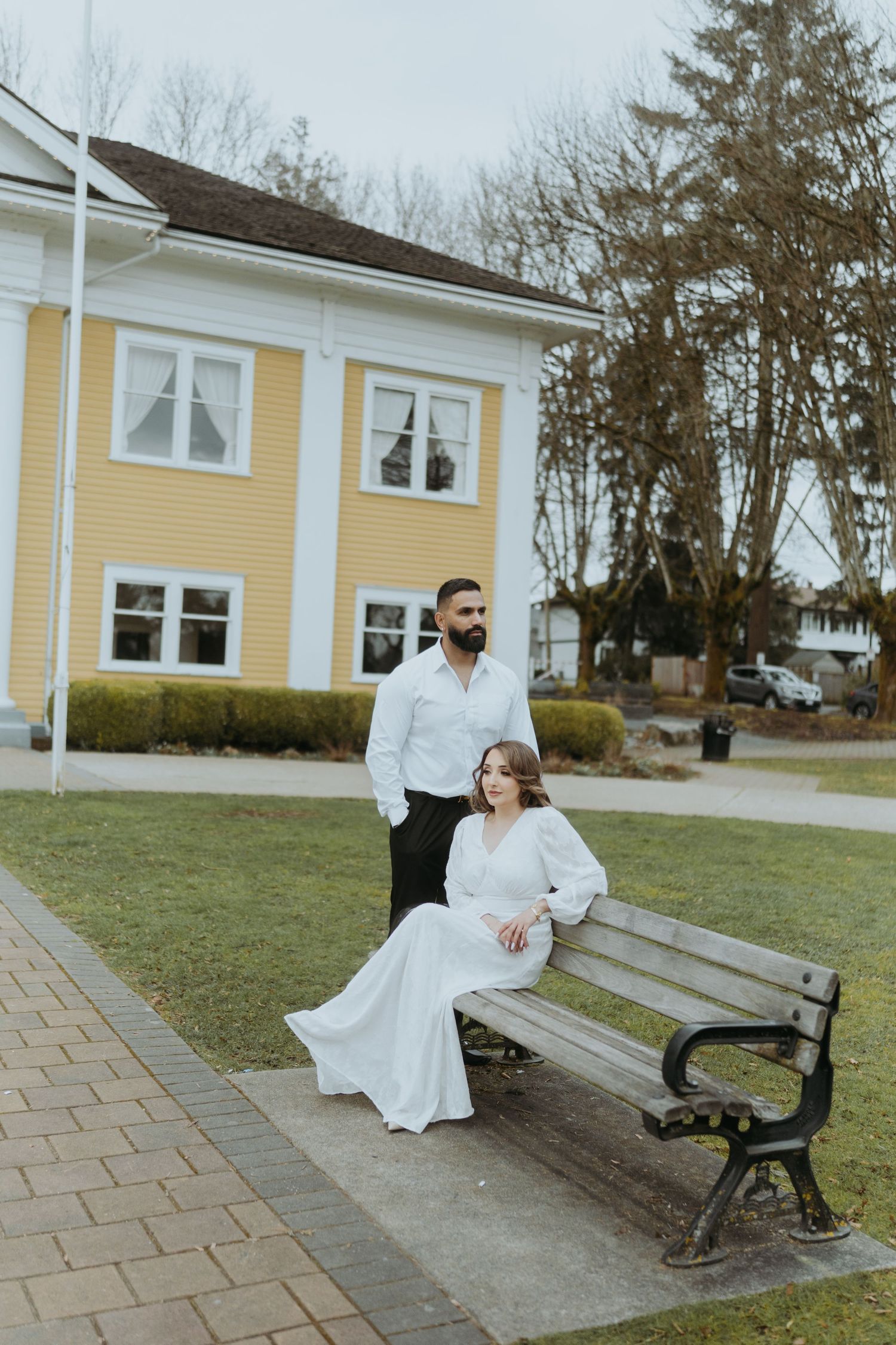 A couple poses together near a bench outside a yellow colonial style building.