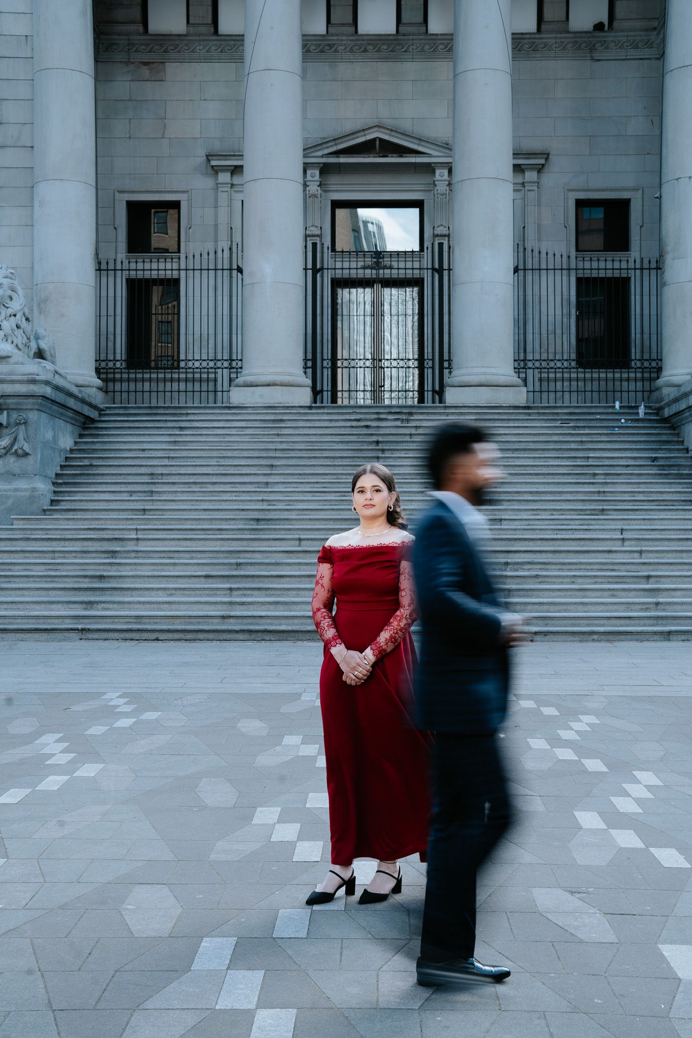 Woman in red off-shoulder gown standing before neoclassical building steps with blurred man passing.