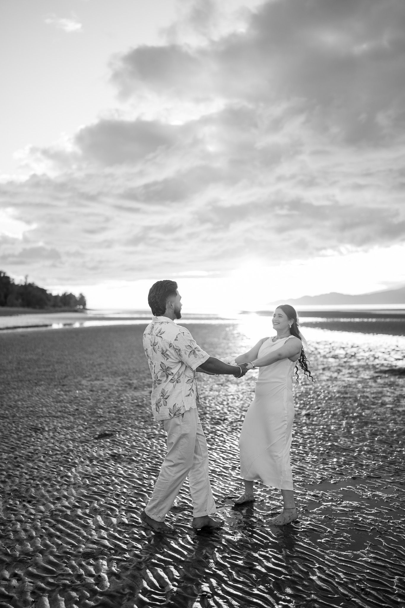 A couple holds hands facing each other on a reflective sandy beach at sunset in black and white.