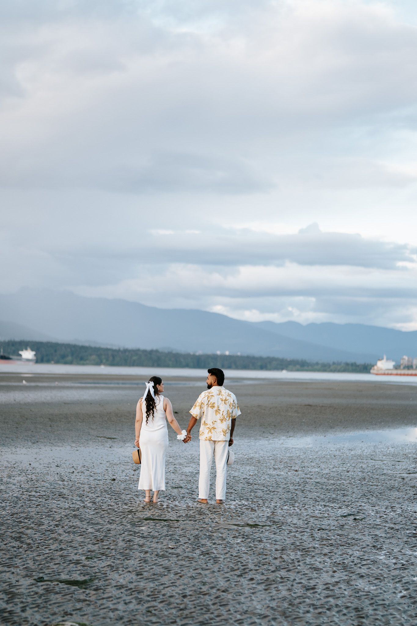 A couple holds hands walking on a sandy beach with mountains and ships visible in the cloudy distance.