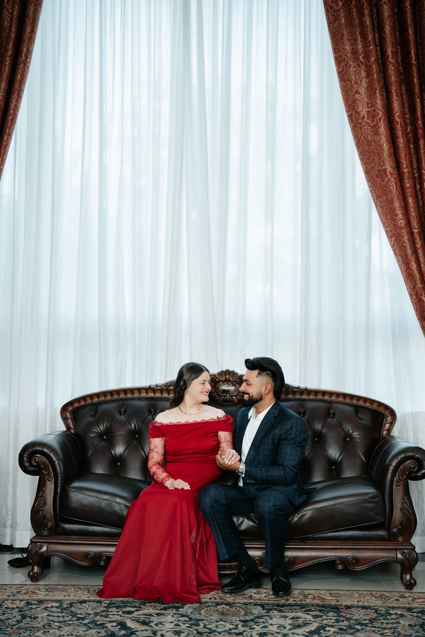 Couple in elegant attire sitting on a brown leather sofa, woman in red gown, man in dark suit, facing each other.