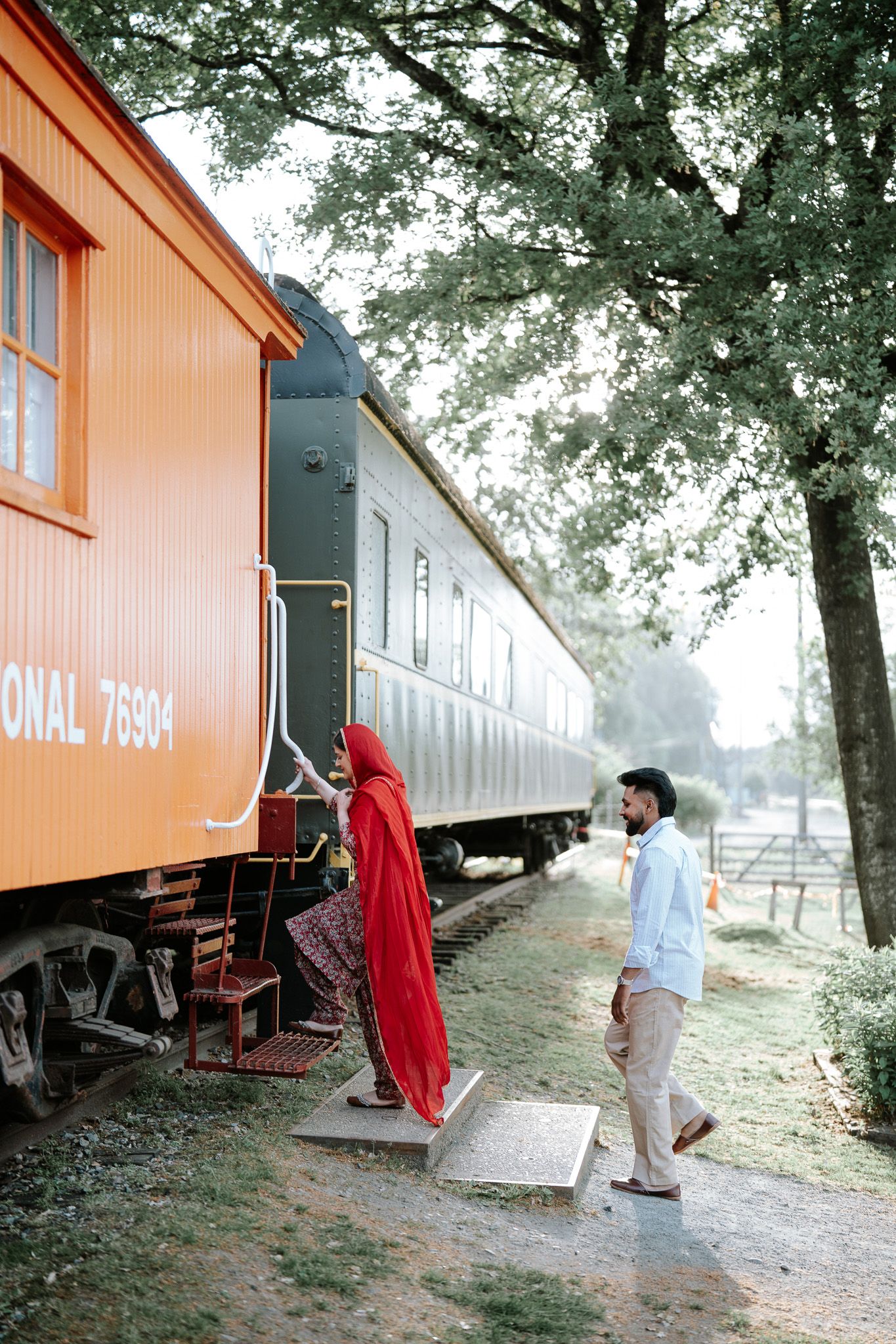Woman in red dupatta boarding vintage orange Canadian National train car as man watches at Fort Langley.