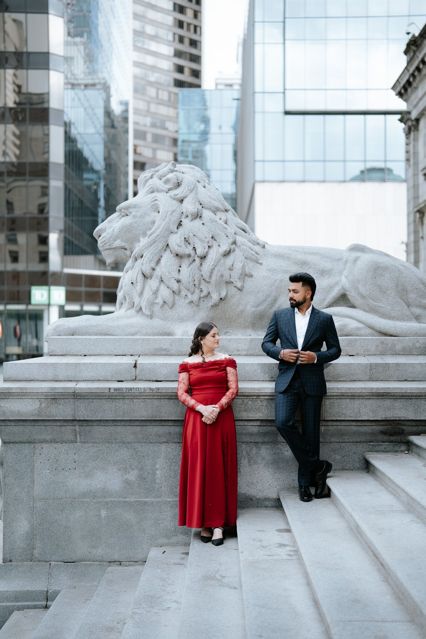 Couple posing beside a stone lion sculpture at a grand city library, woman in red gown, man in dark suit.
