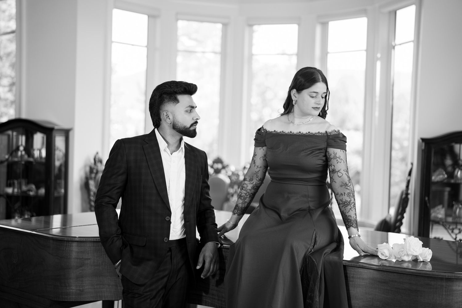 Black and white photo of an elegant couple standing by a grand piano in a bright windowed room.