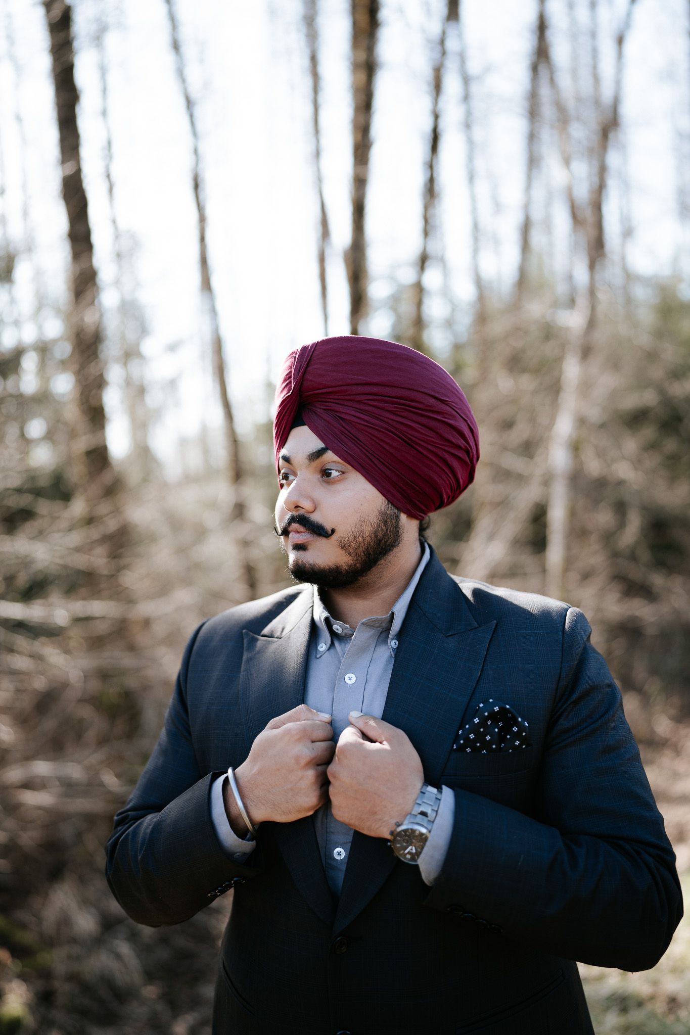 A man wearing a maroon turban and dark suit adjusts his collar in a series of three poses against a wooded background.