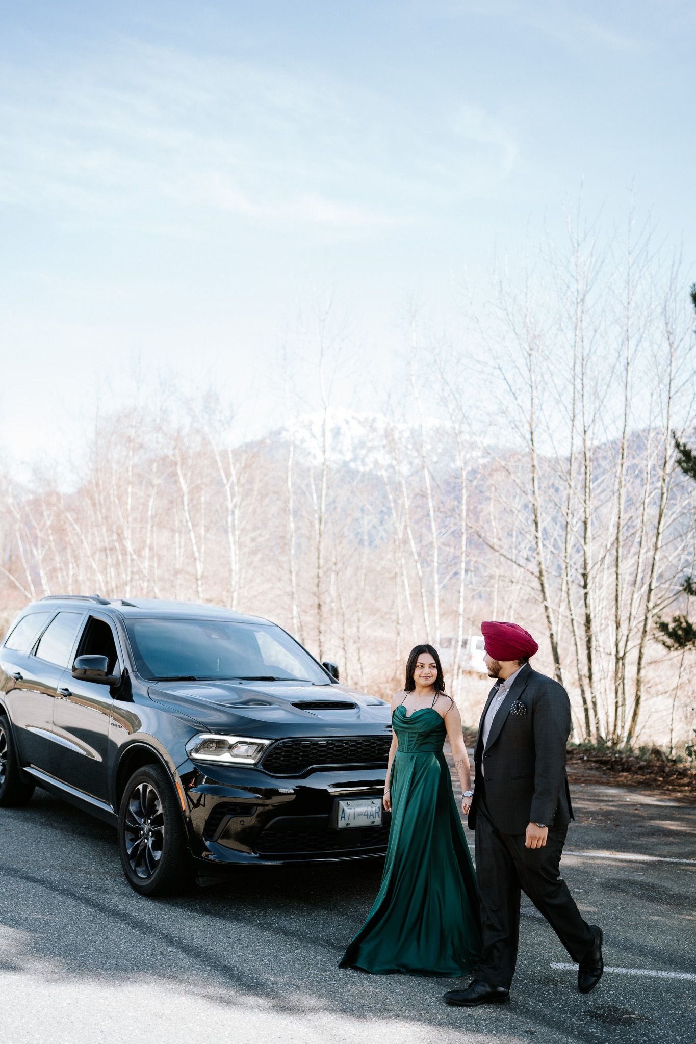 A couple in formal attire poses next to a black SUV on a winter road with bare trees in the background.