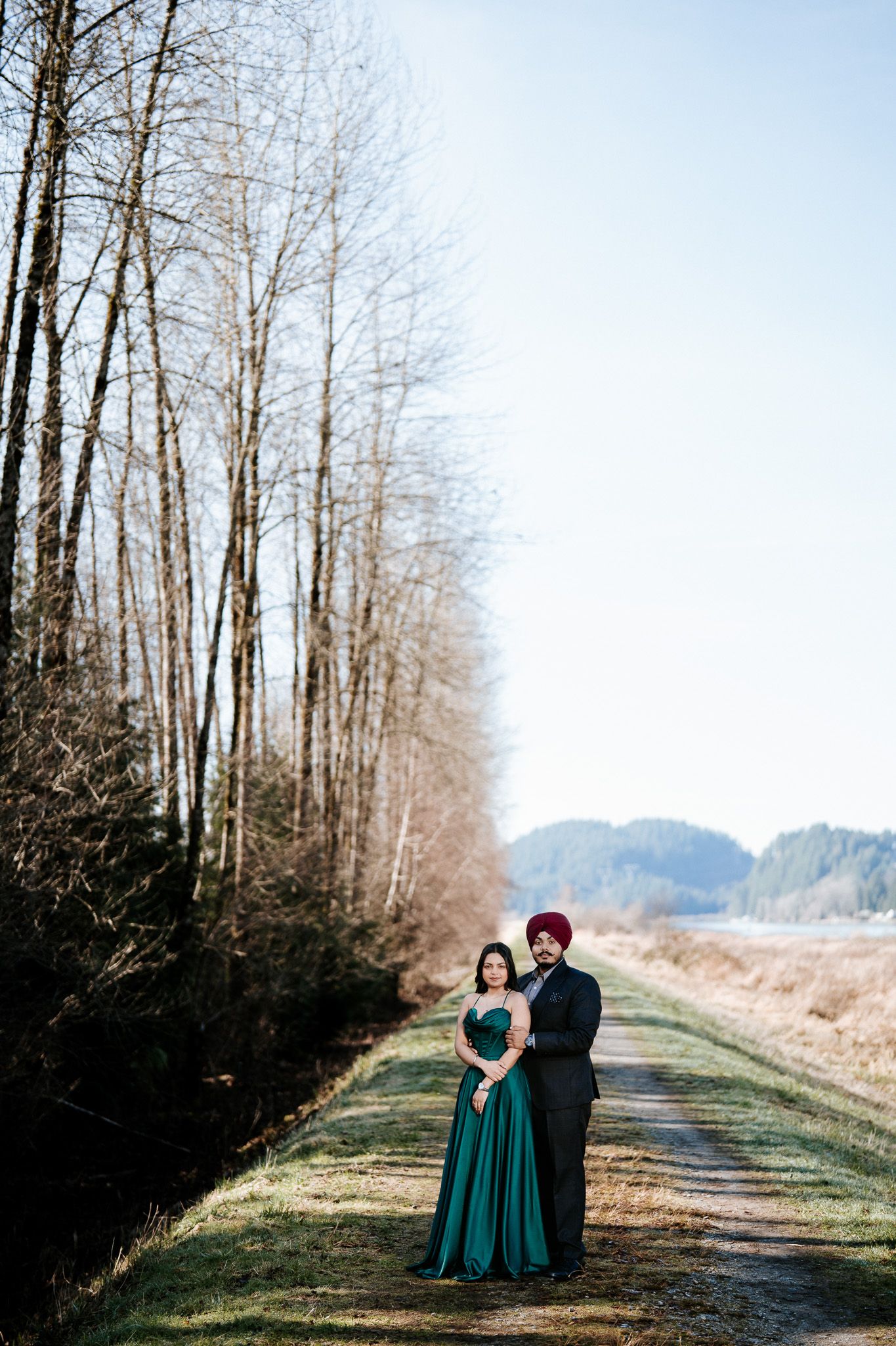 A couple in formal attire stands on a tree-lined path during a winter photo session in nature.