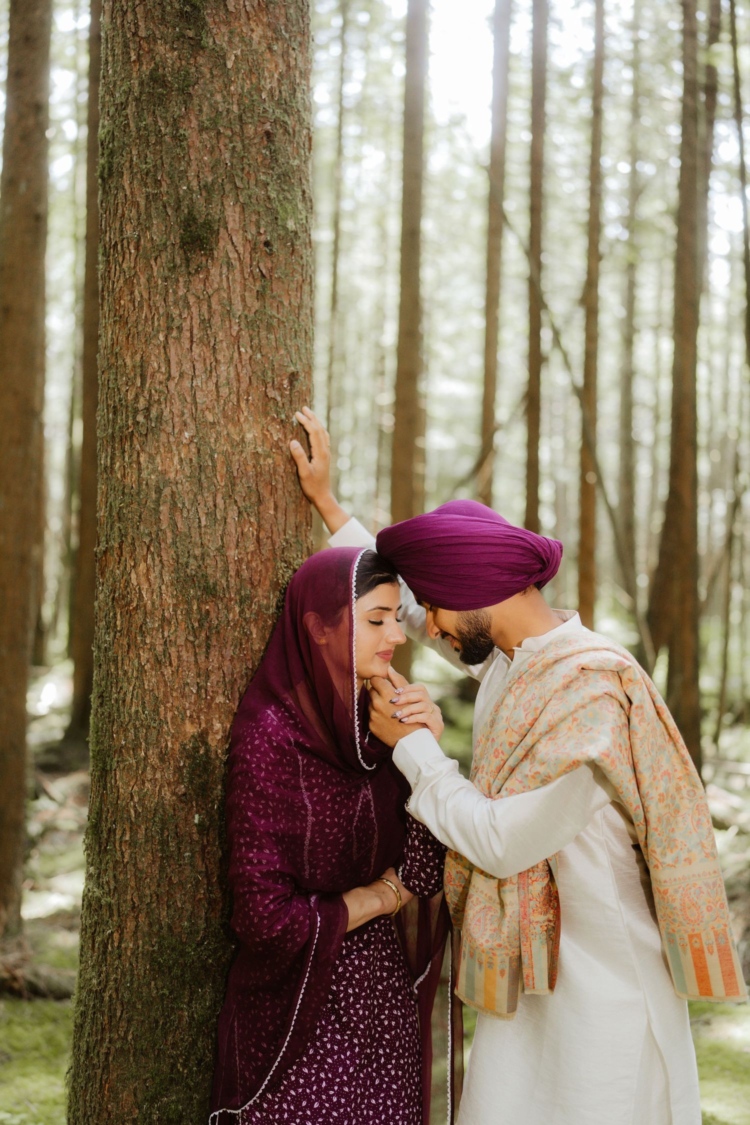A Sikh couple in traditional attire shares a romantic moment leaning against a tree in a forest.