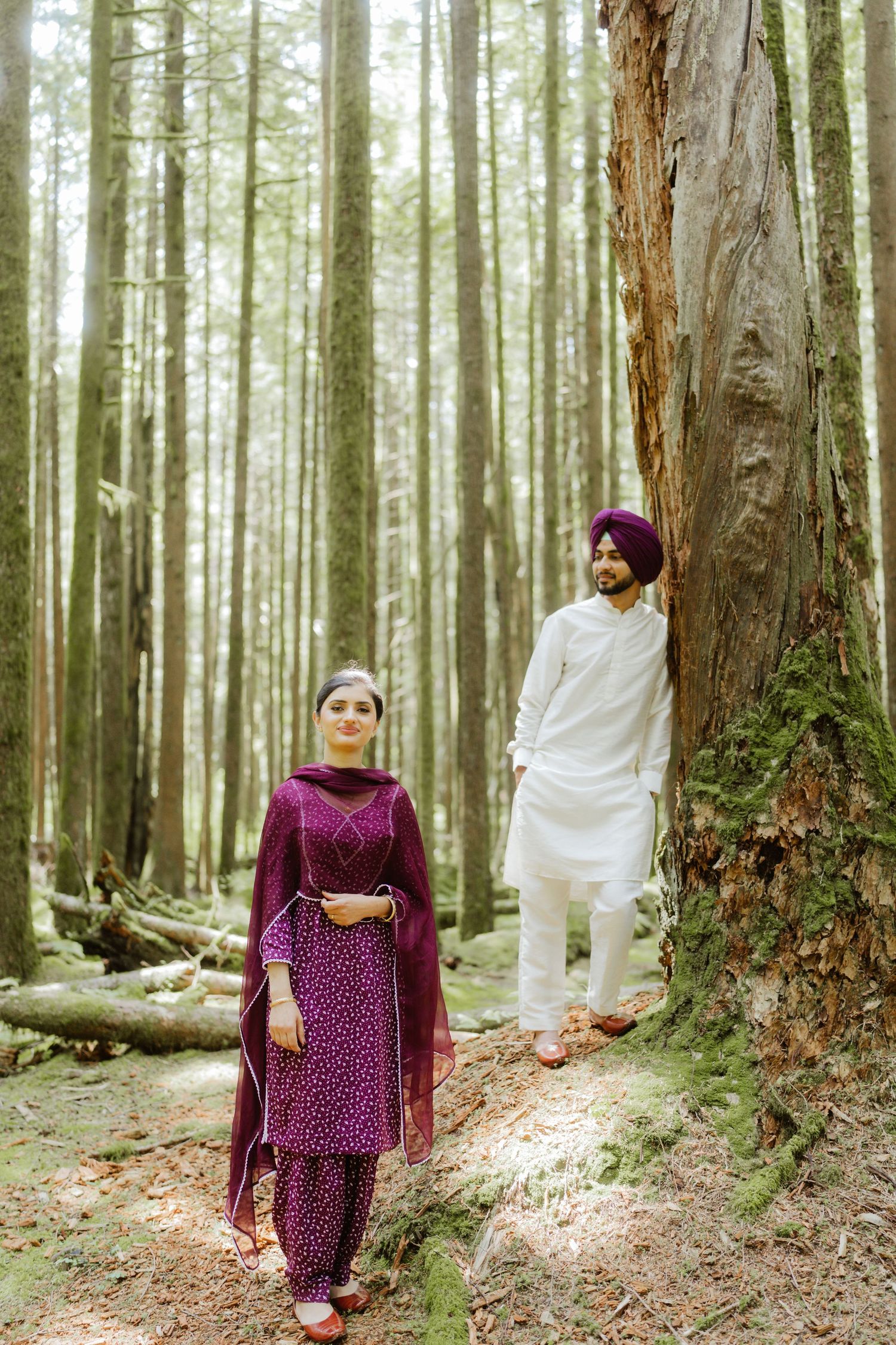 Sikh couple in traditional attire posing in a tall forest, woman in purple salwar kameez, man in white with orange turban.