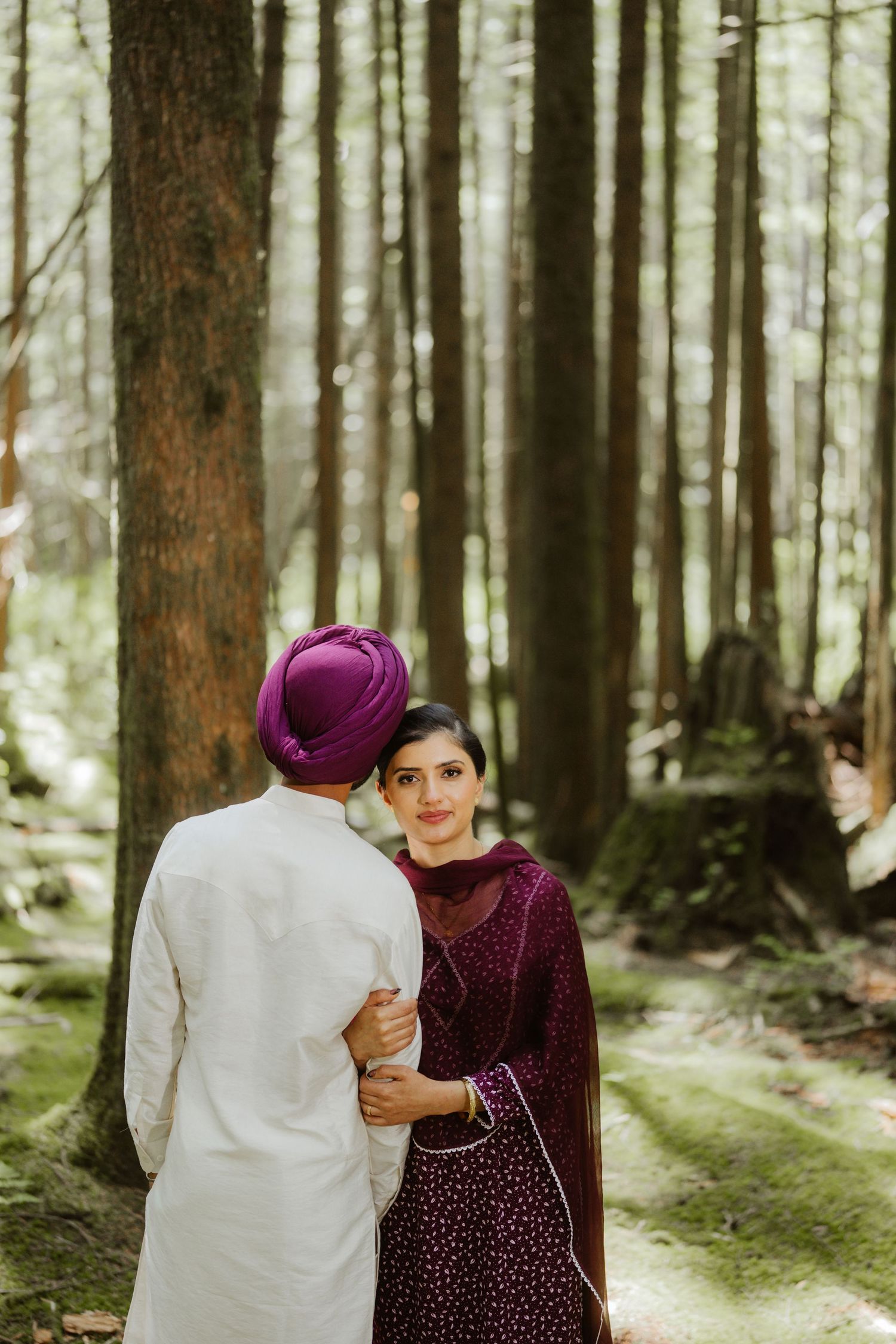 Sikh bride in maroon salwar smiling at groom in white kurta in forest.