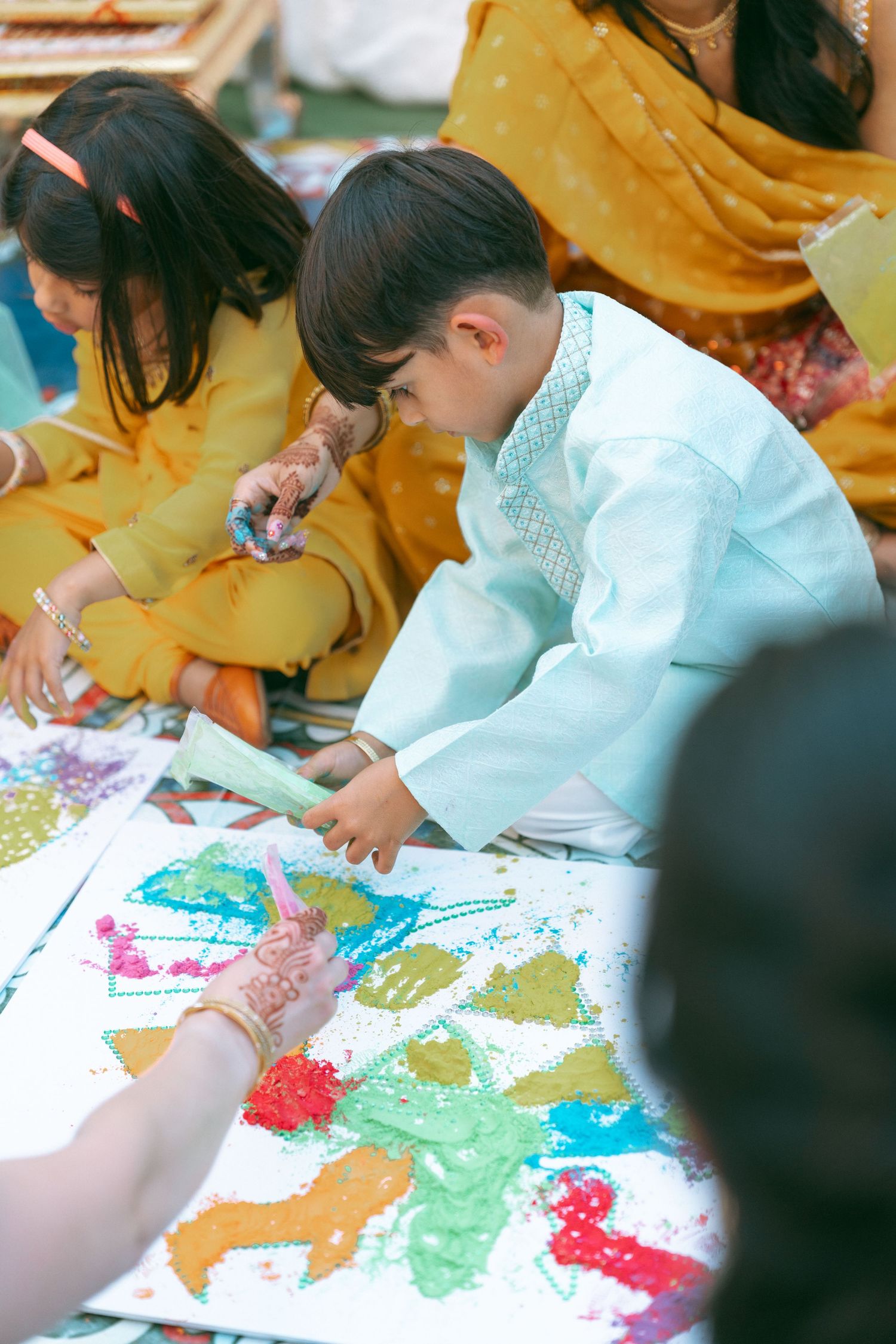 Children in traditional attire painting colorful artwork on white canvas during a cultural celebration event.
