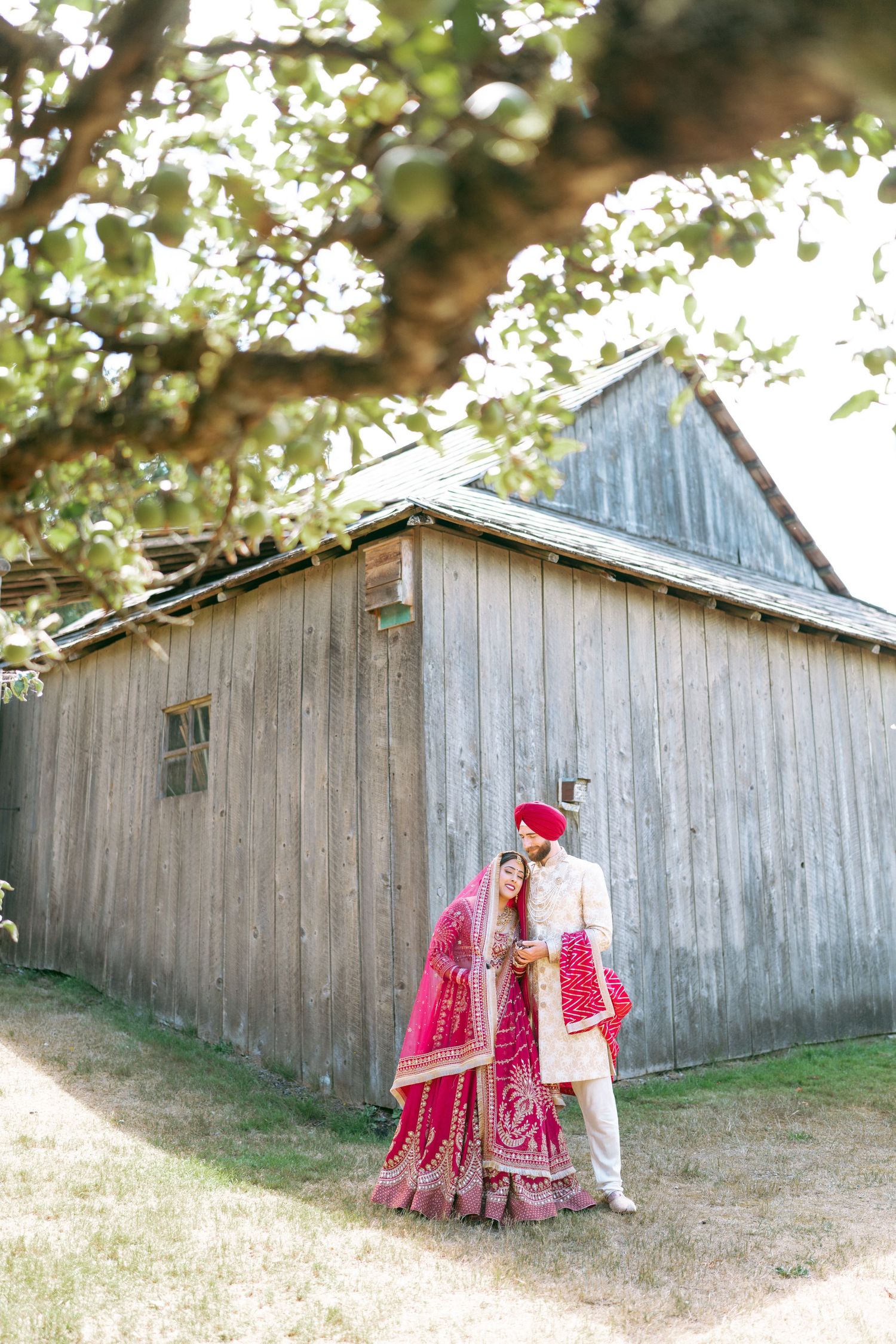 Indian bride and groom in traditional red and gold wedding attire pose romantically beside a rustic wooden barn.