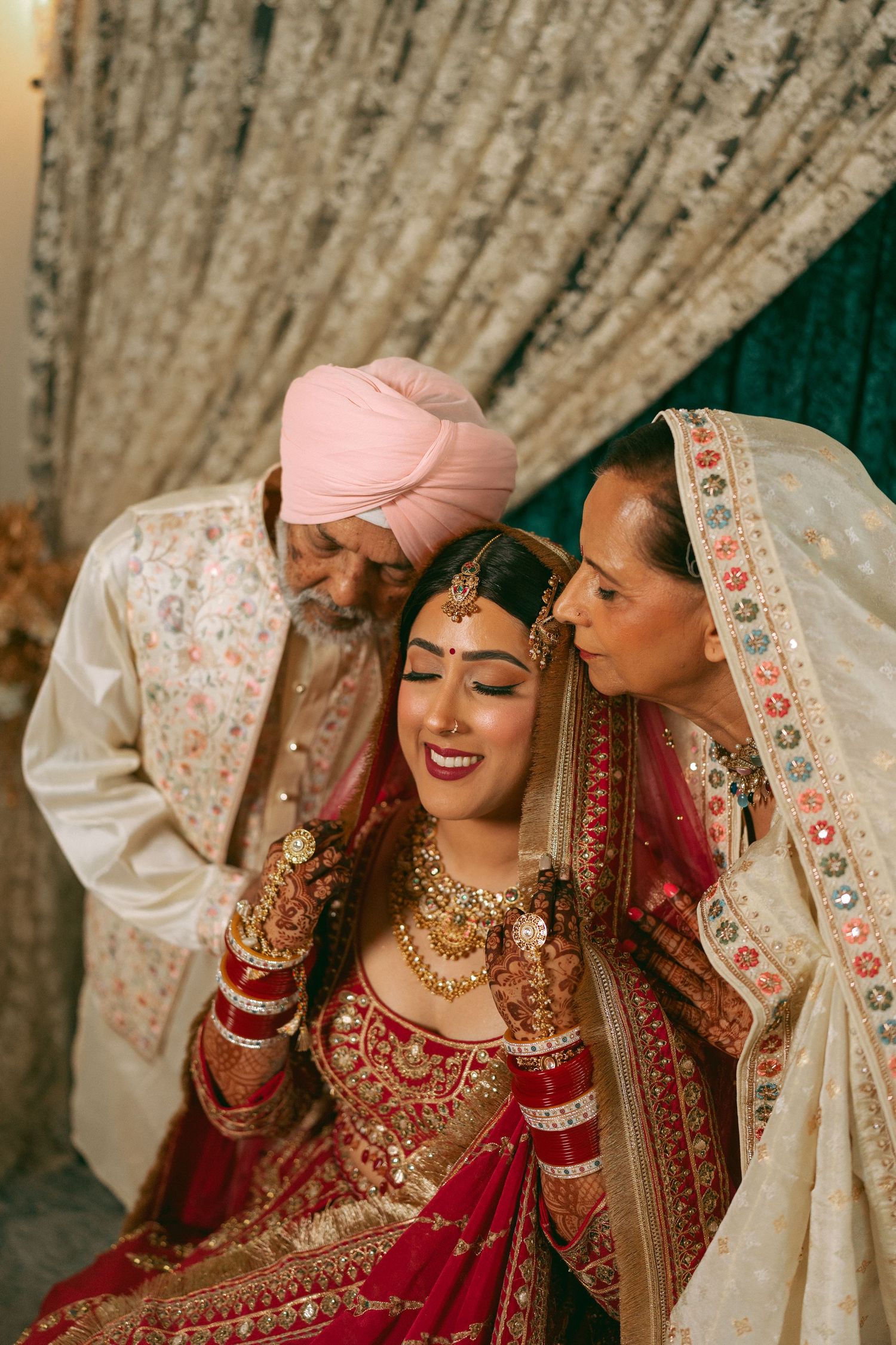 Sikh bride in red lehenga with gold jewelry flanked by parents in traditional attire at wedding ceremony.