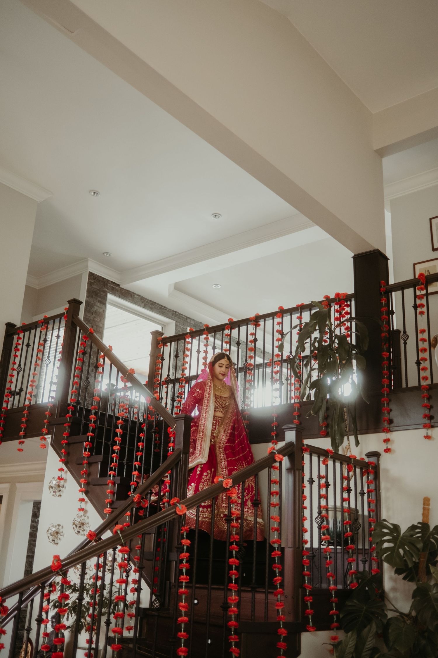 Bride in red bridal lehenga standing on decorated staircase with red floral garlands at South Asian wedding.