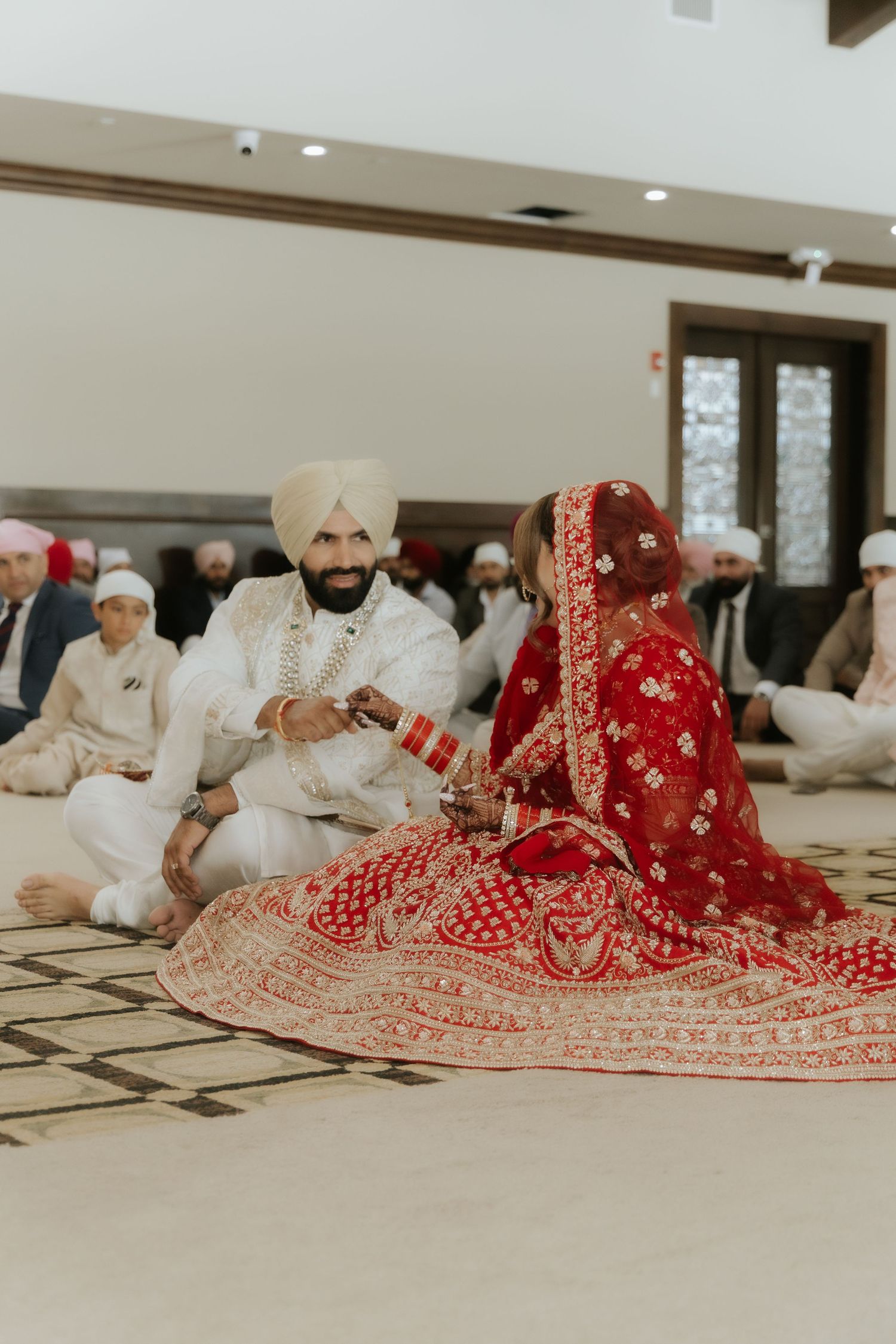 Sikh bride in red lehenga and groom in white sherwani and turban during Anand Karaj wedding ceremony.