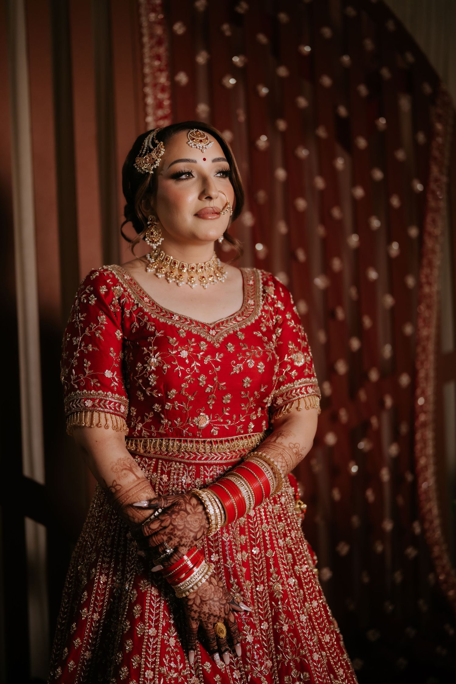 Indian bride in red lehenga with gold embroidery and traditional jewelry poses elegantly for bridal portrait.
